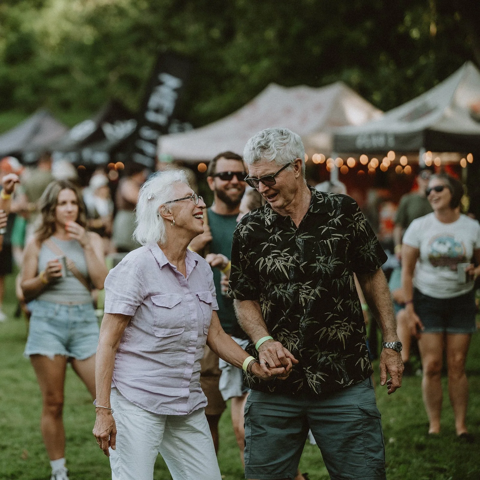 A joyful elderly woman and man are dancing and holding hands at an outdoor event with tents and string lights, surrounded by people.