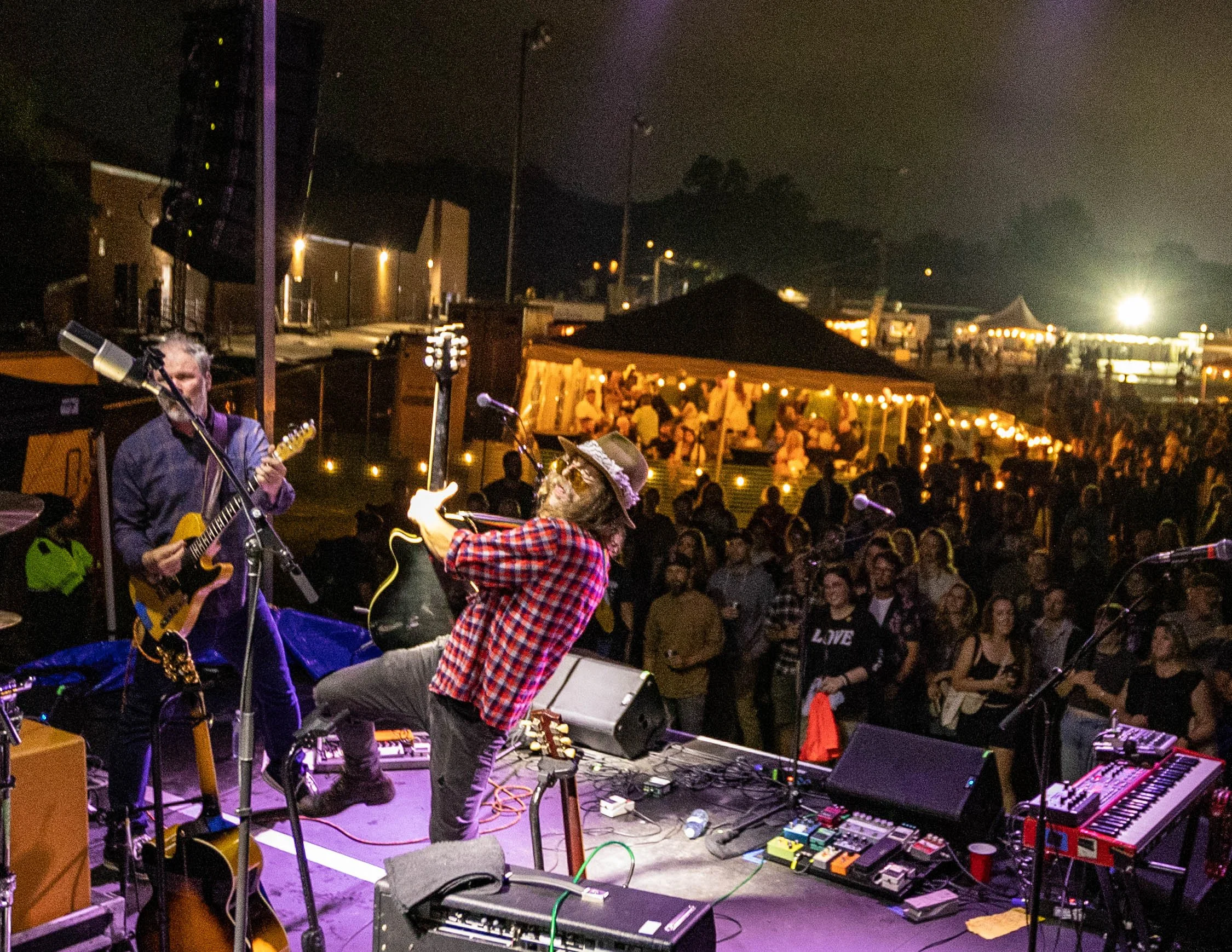 A live outdoor music performance at night with two musicians on stage: one playing guitar and another playing a guitar while leaning back. A crowd is watching the show, with tents and string lights visible in the background.