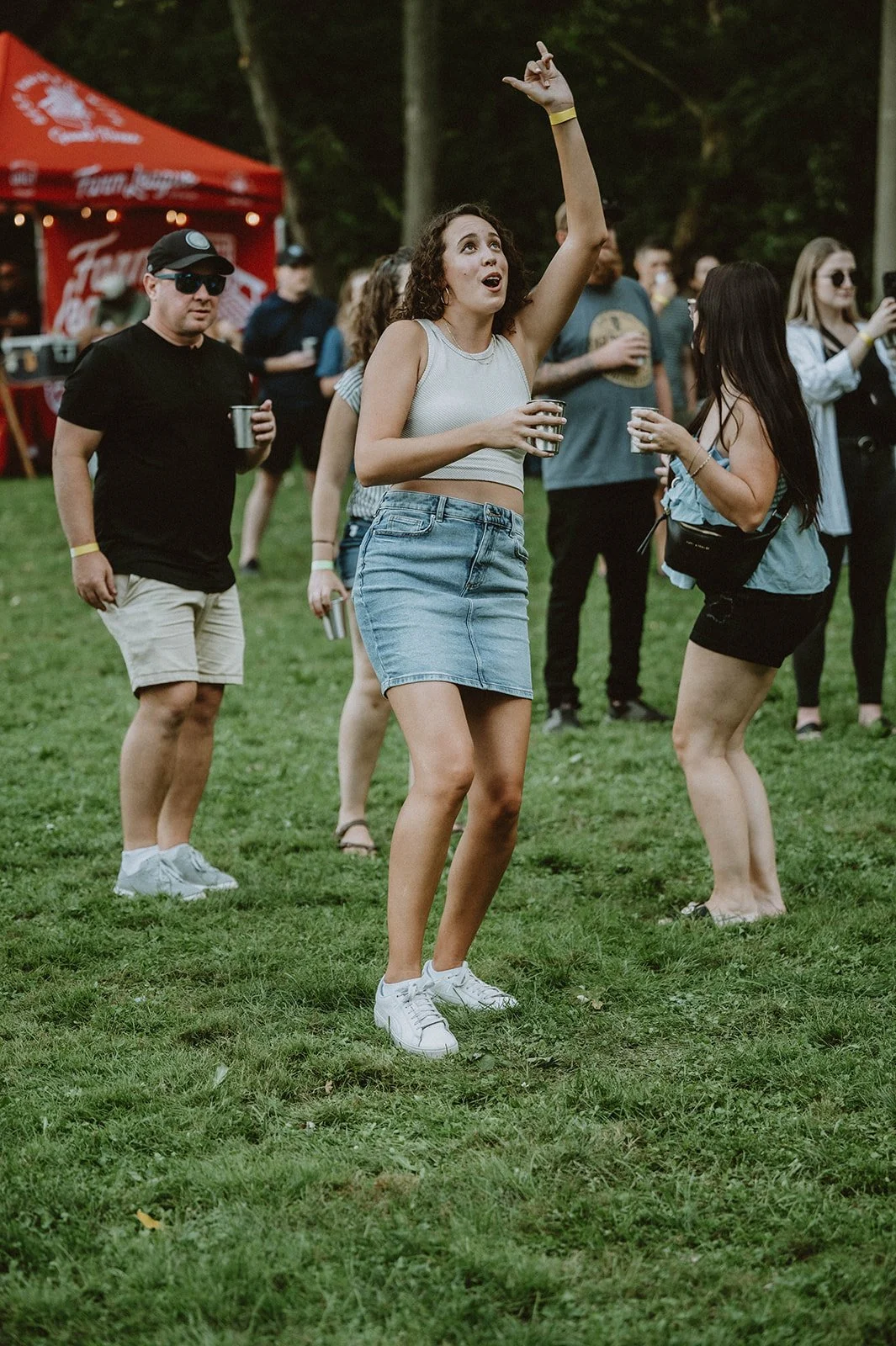 People enjoying a music festival outdoors, with a woman dancing and raising her hand, others holding drinks, and a red tent in the background.
