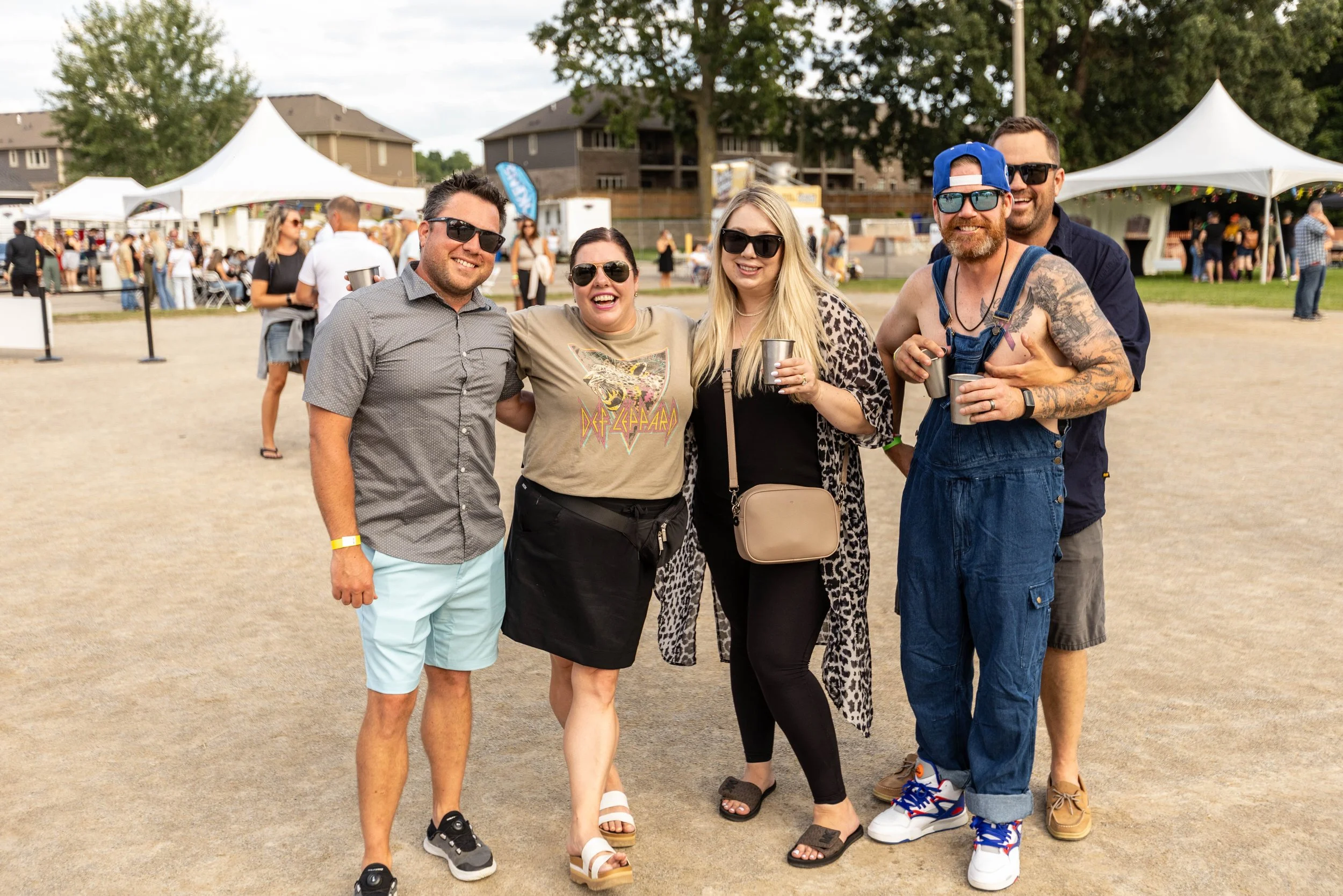 Group of six friends enjoying an outdoor festival, standing on a dirt field with tents and other festival-goers in the background, smiling at the camera.