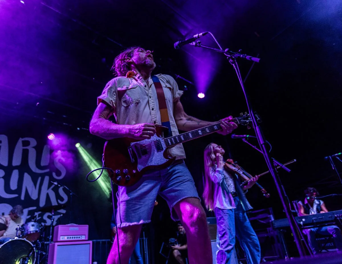 A live music performance with a male guitarist in the foreground, wearing a beige shirt and shorts, with long hair and a beard, playing electric guitar on stage under purple and green lights. Other band members are seen in the background with instrum