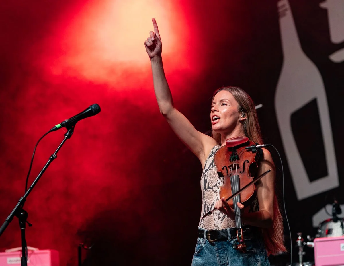 A woman performing on stage, holding a violin and raising her right arm, with a microphone in front of her and stage equipment visible in the background, illuminated by red stage lighting.