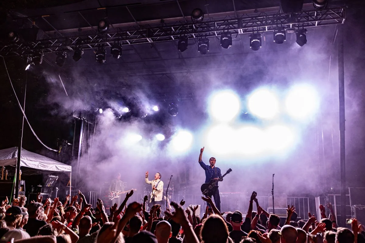 A live outdoor concert at night with a band performing on stage illuminated by bright white lights, fog effects, and an enthusiastic crowd with raised hands.