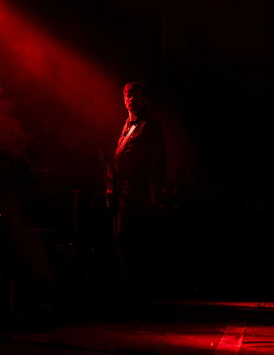 A person standing on stage illuminated by red lighting, holding a guitar, with a dark background.