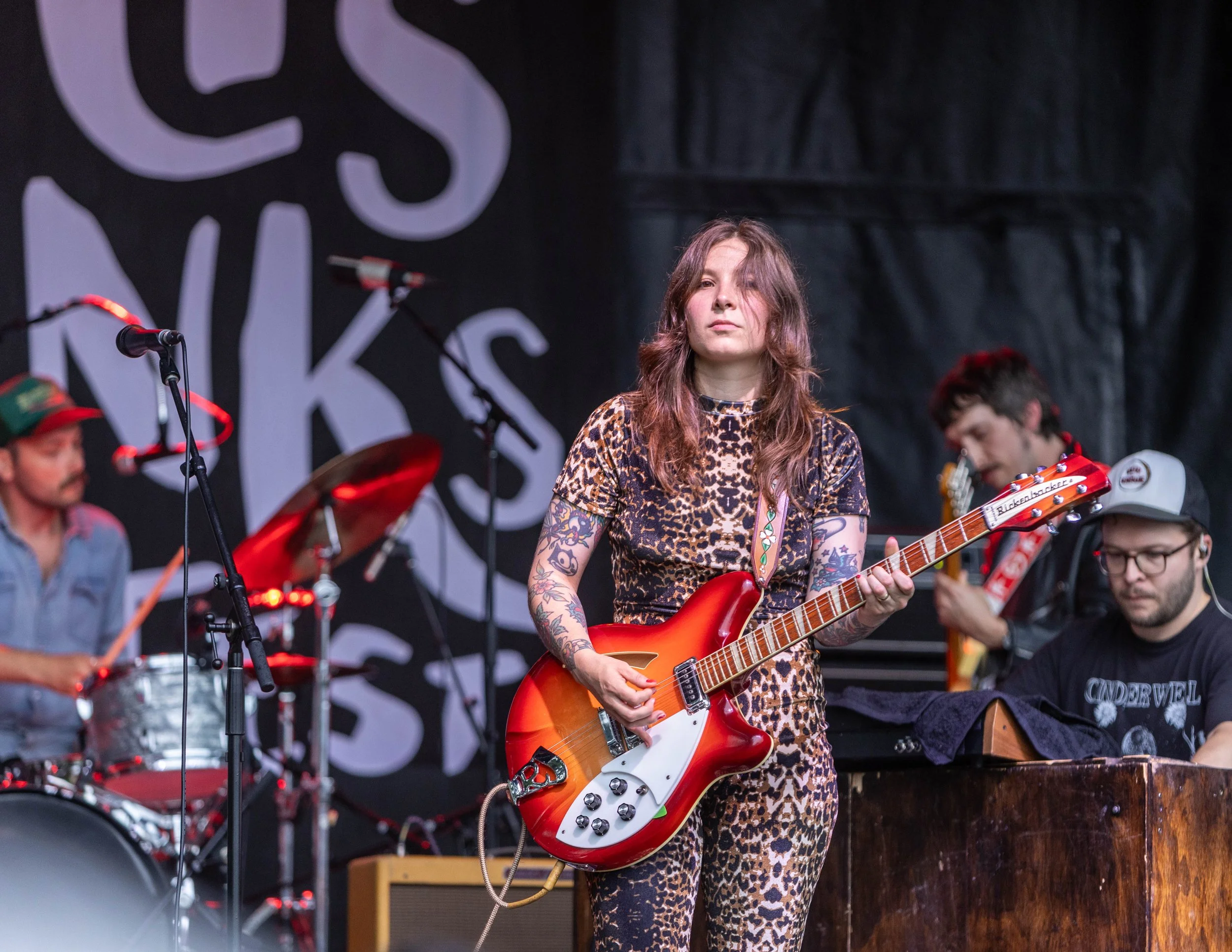 A woman playing an orange and white electric guitar on stage with a band, wearing a leopard print outfit, with tattoos on her arms, during a live music performance.