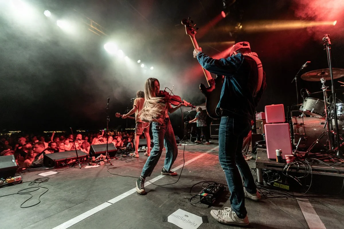 Musicians performing on stage with a crowd watching, including a violinist and a bassist, under concert lights.
