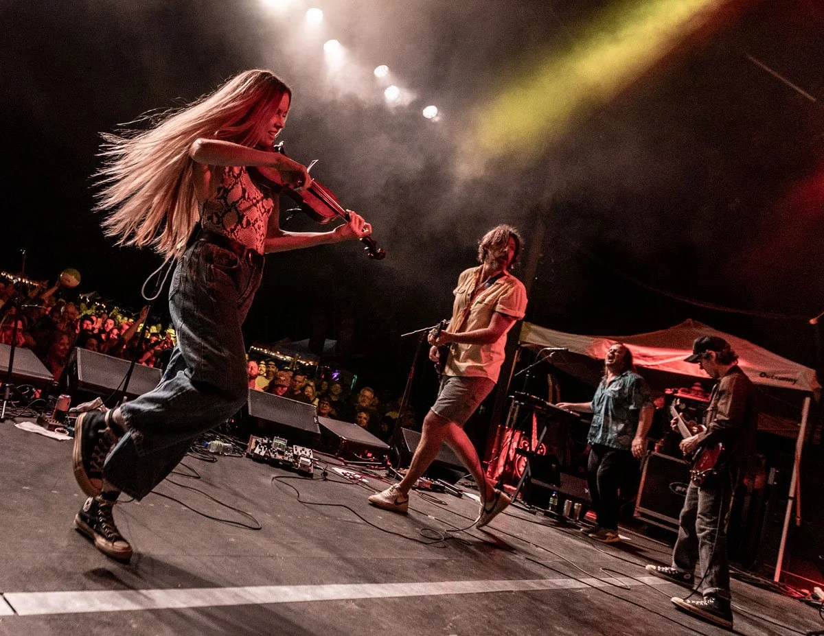 Musicians performing on stage at a concert with red and yellow lighting and an audience in the background.