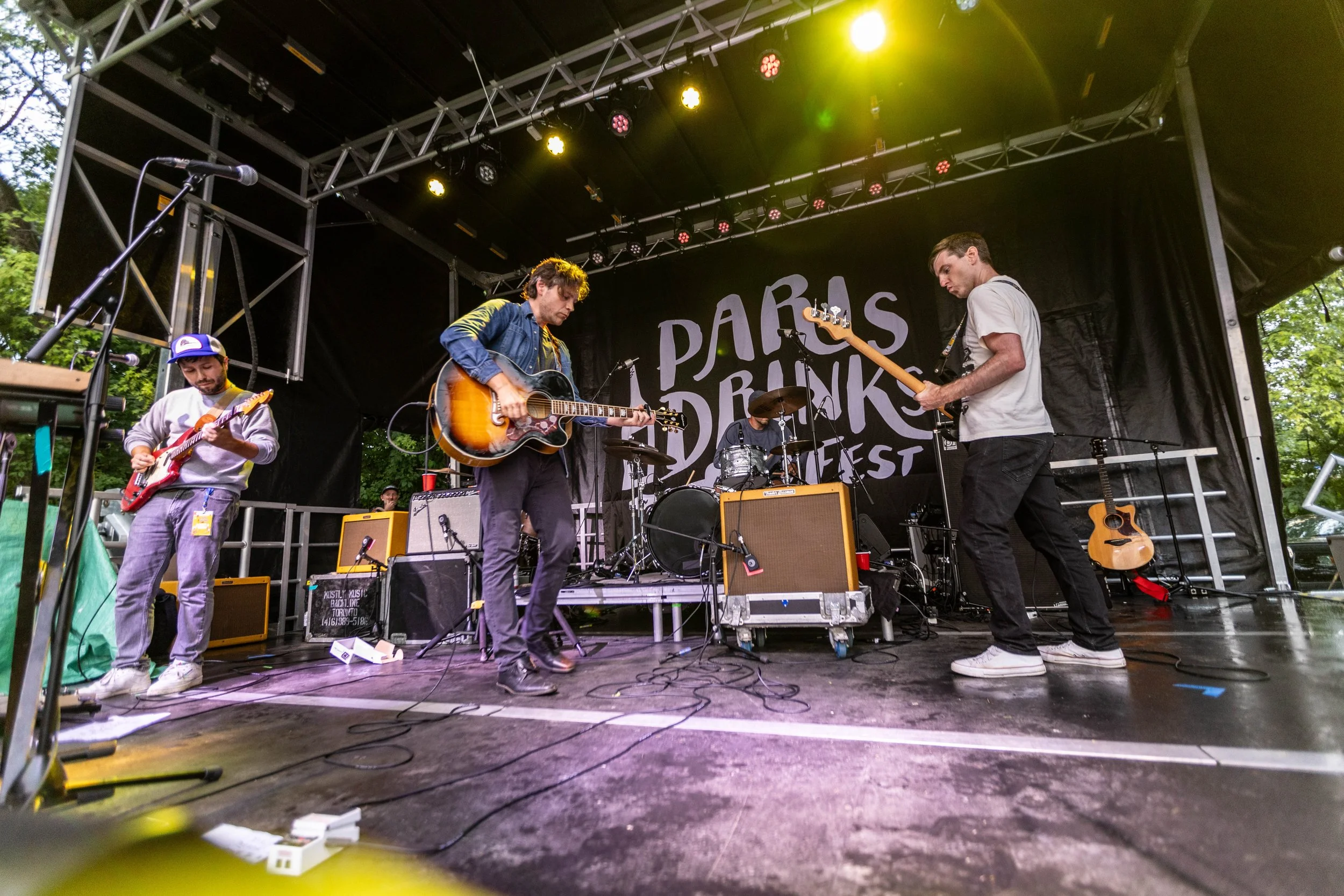 A band of four musicians performing on a stage outdoors at a music festival, with a backdrop reading 'París Drinks Fest' and trees in the background.