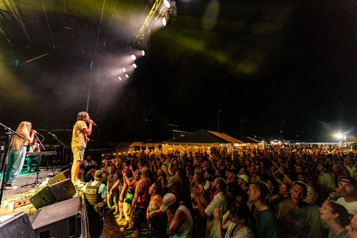 Crowd at an outdoor concert at night, stage with performers, large audience, tents, and bright lights.