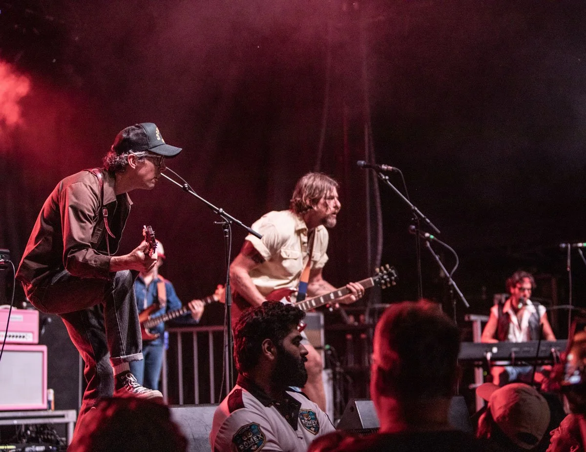 A band performing on stage with four musicians playing guitars and keyboard, under red stage lighting, in front of an audience.