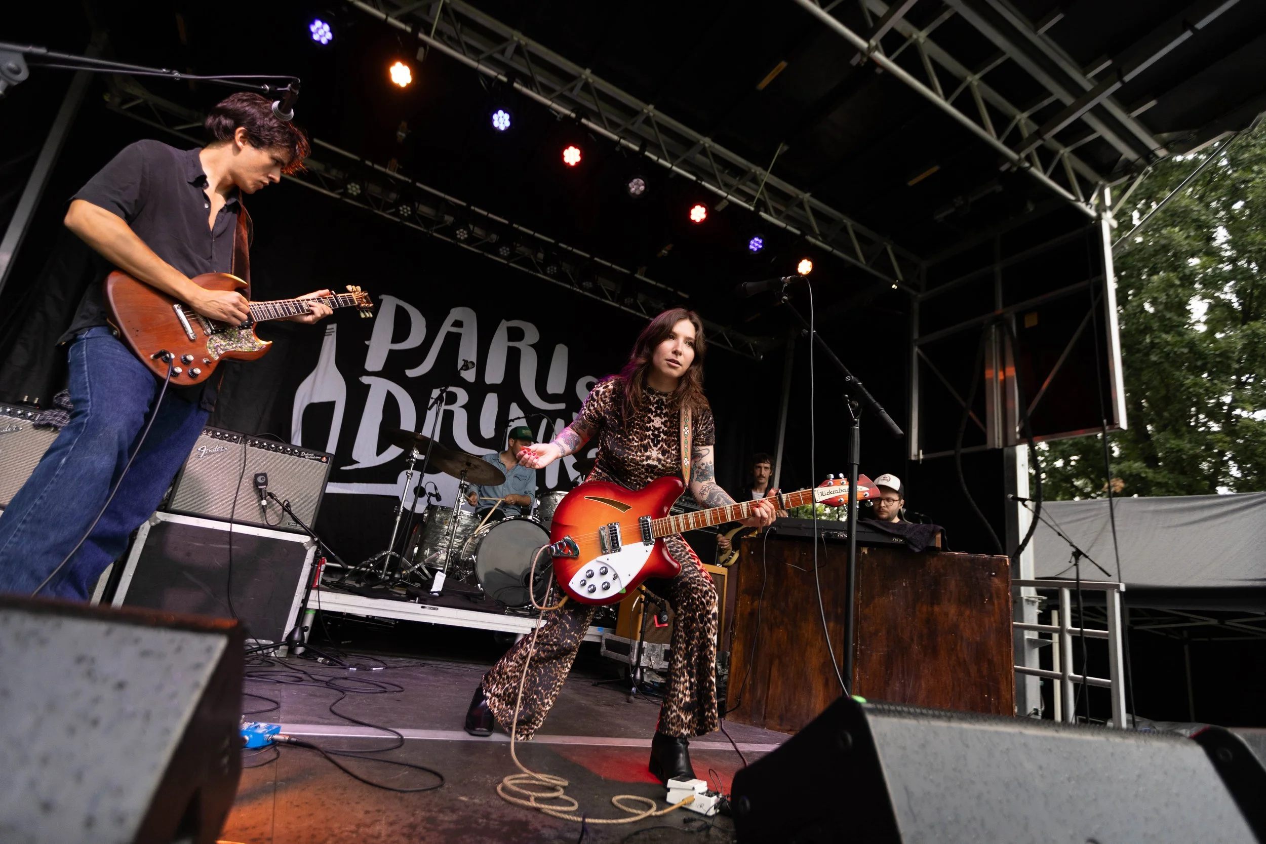 A band performing on stage at an outdoor music festival with a woman in leopard print pants and a leopard print top playing guitar in the center. On the left, a man is playing a guitar, and in the background, a drummer is visible. The backdrop has th