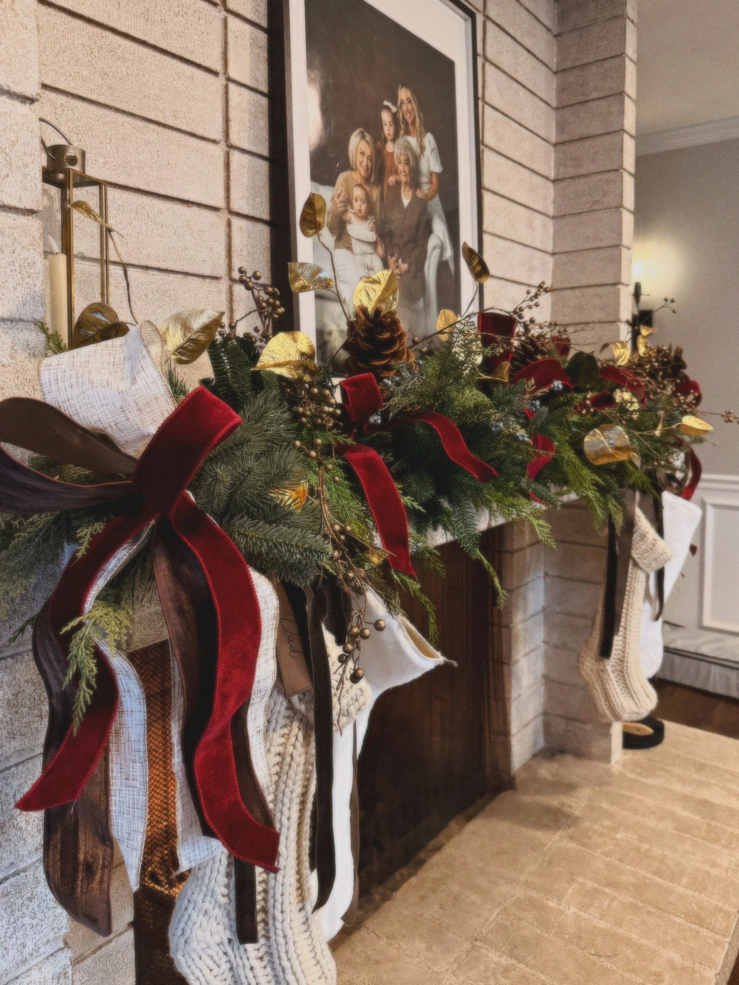 Decorative Christmas garland with stockings hanging from a fireplace, adorned with red ribbons, gold leaves, pinecones, and greenery, in front of a brick wall with a family portrait.