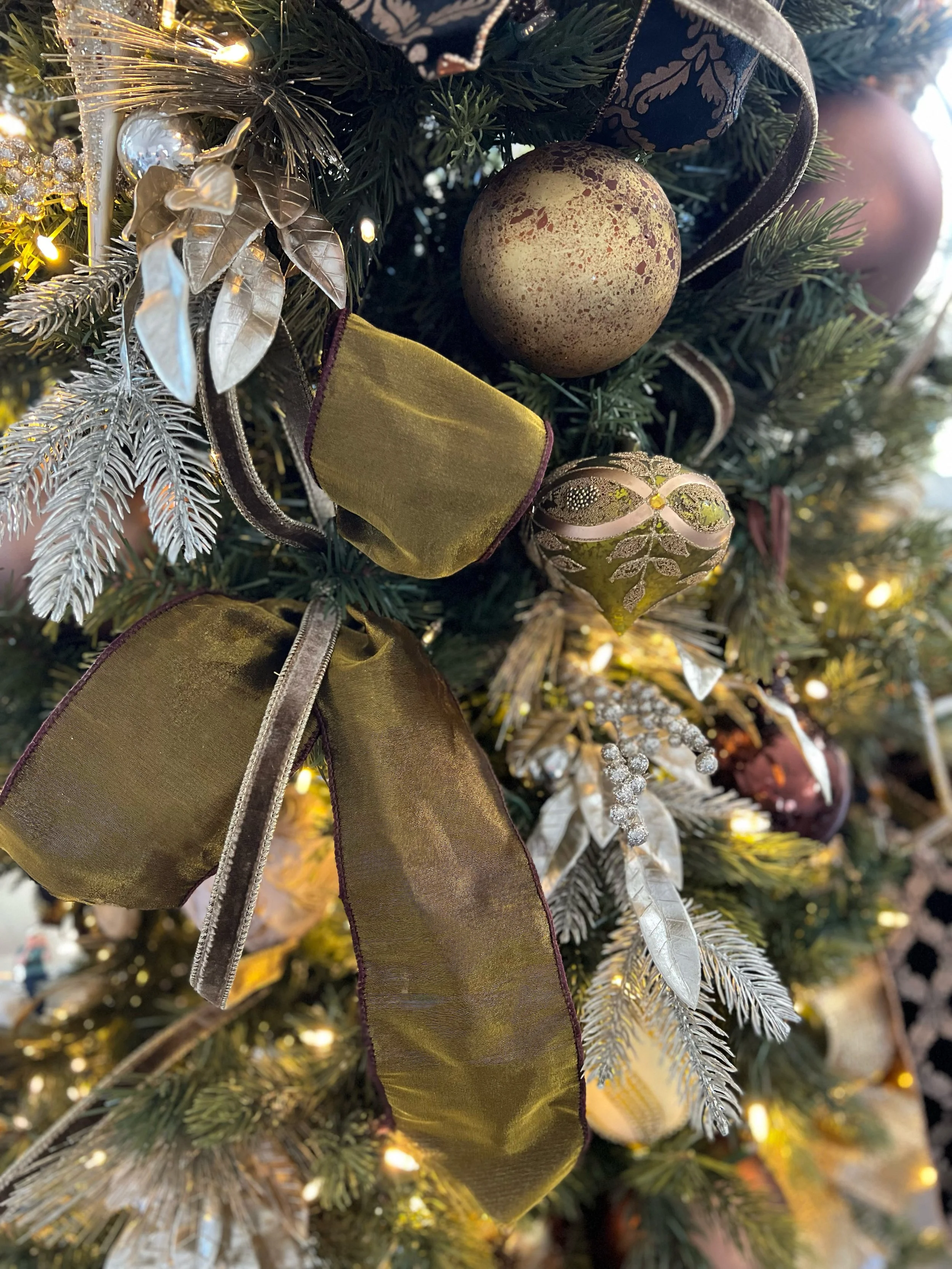 Close-up of a Christmas tree decorated with gold and silver ornaments, ribbons, and white and silver leaves, with warm white lights.