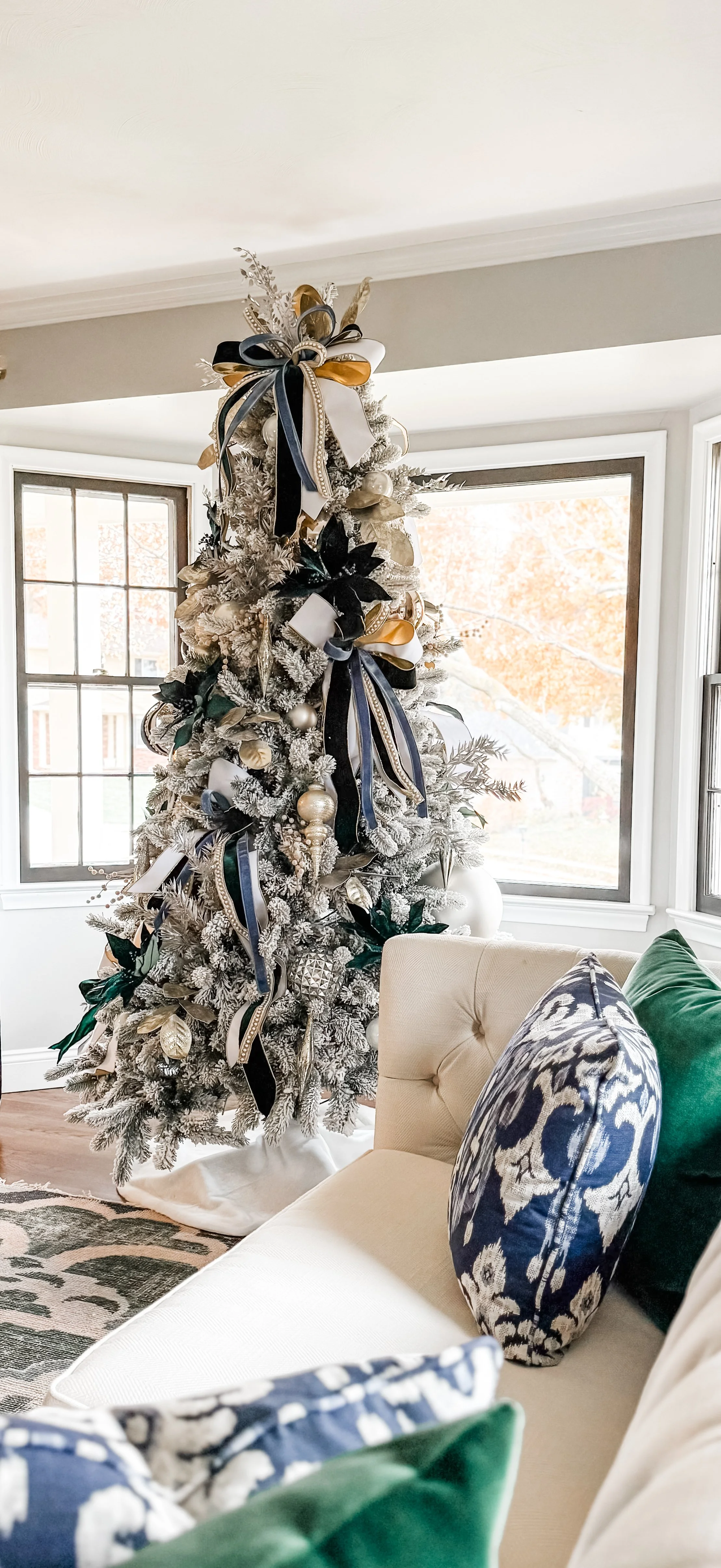 A decorated white Christmas tree with ribbons, ornaments, and faux snow, placed in a well-lit living room with large windows and a beige sofa with patterned and green pillows.
