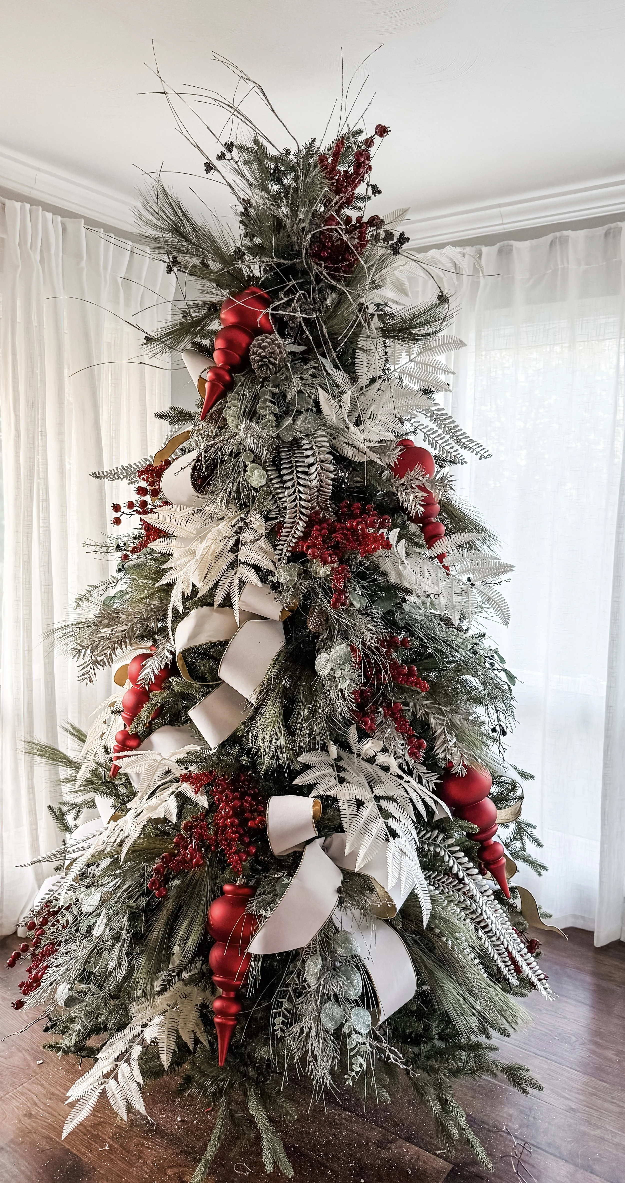 Decorated Christmas tree with red, white, and gold ornaments, white ribbons, and various green and white foliage against a background of white curtains and wooden floor.