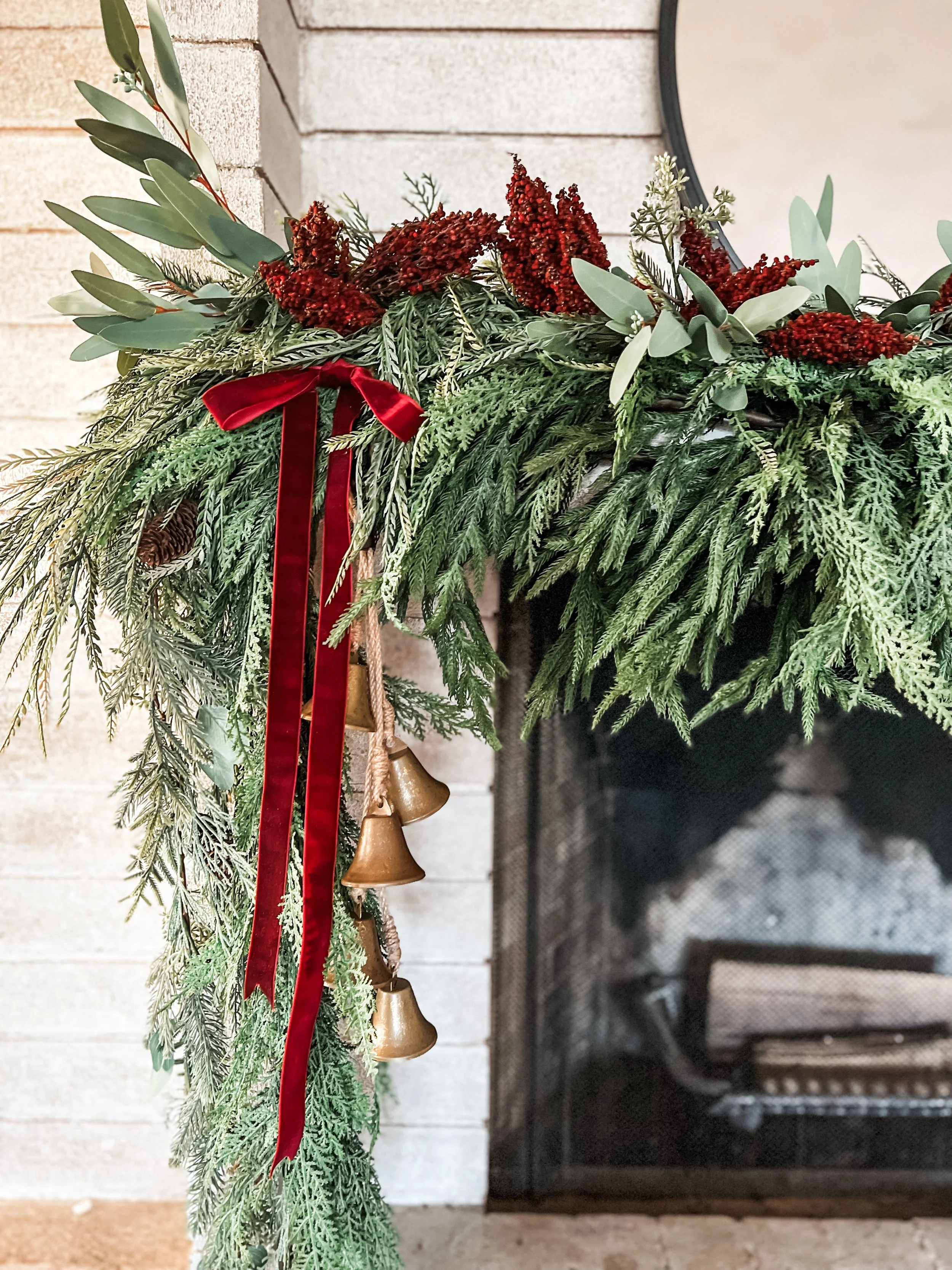 Close-up of holiday greenery and decorations on a fireplace mantel, including pine branches, red berries, leaves, a red ribbon, and gold bells.
