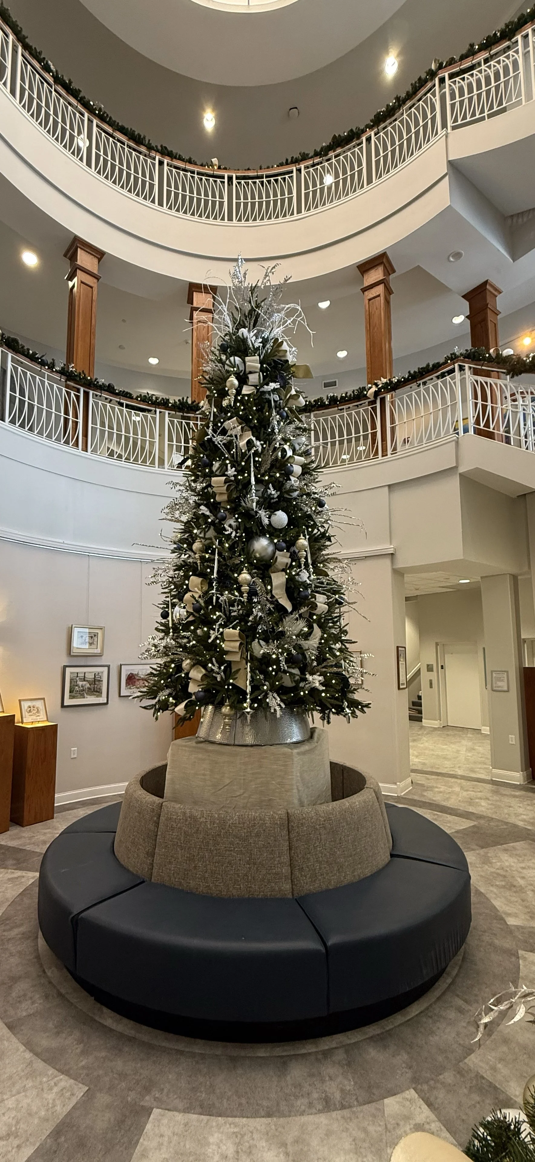 A decorated Christmas tree in the center of a lobby with a circular bench around it, featuring silver, black, and white ornaments and ribbon, surrounded by a modern interior with a curved balcony and framed pictures on the wall.