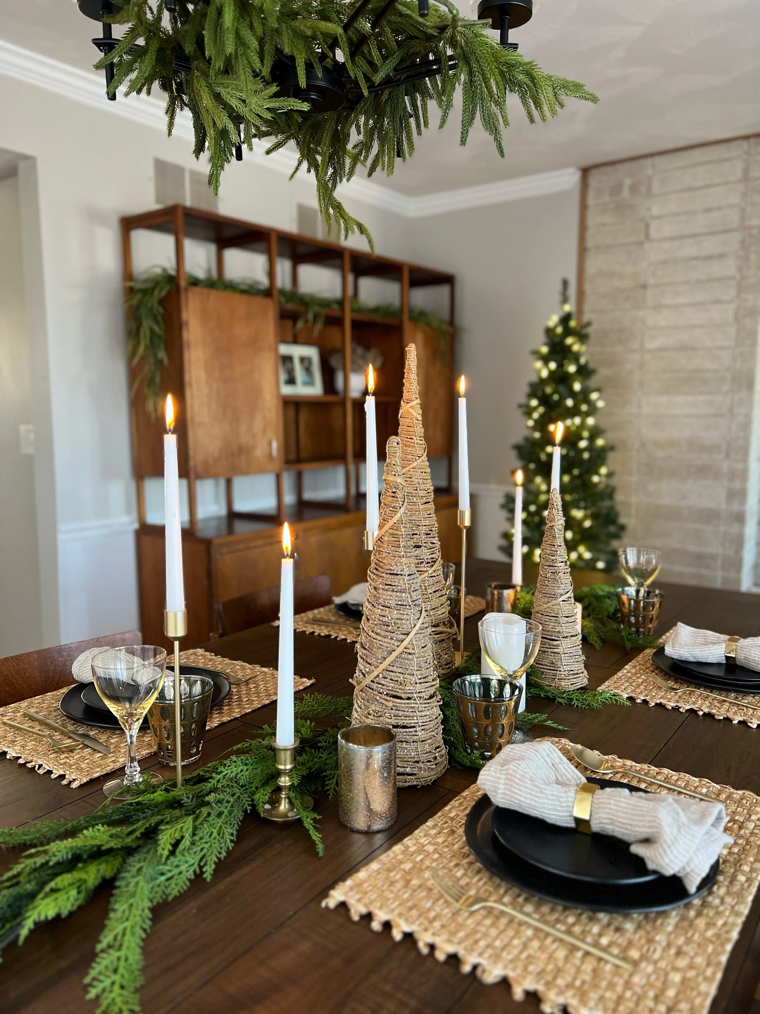 A festive dining table decorated with woven placemats, black dinnerware, white cloth napkins with gold napkin rings, and gold flatware. The centerpiece includes cone-shaped ornaments resembling Christmas trees, topped with candles, surrounded by gree
