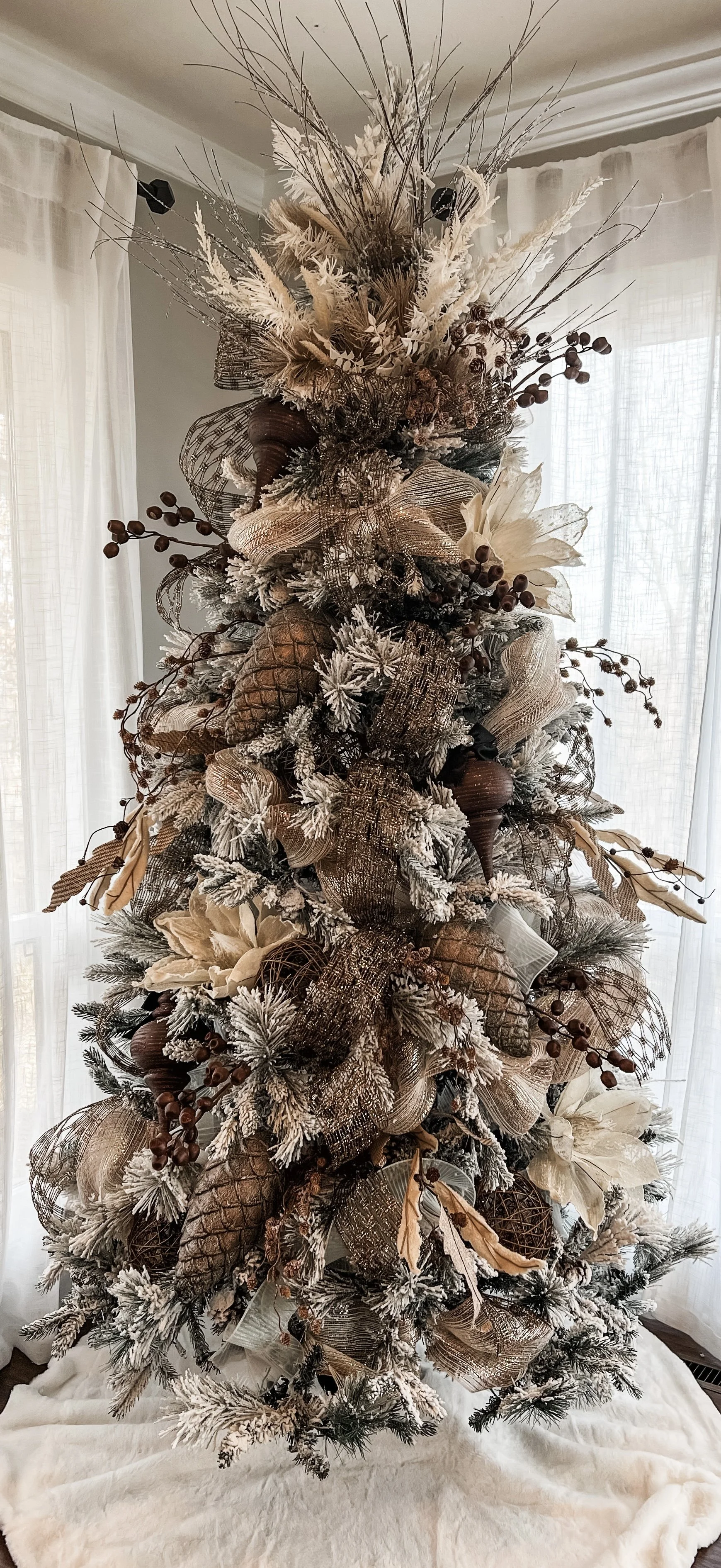 Decorated Christmas tree with gold, brown, and cream ornaments, ribbons, and faux snow, placed near windows with light curtains.