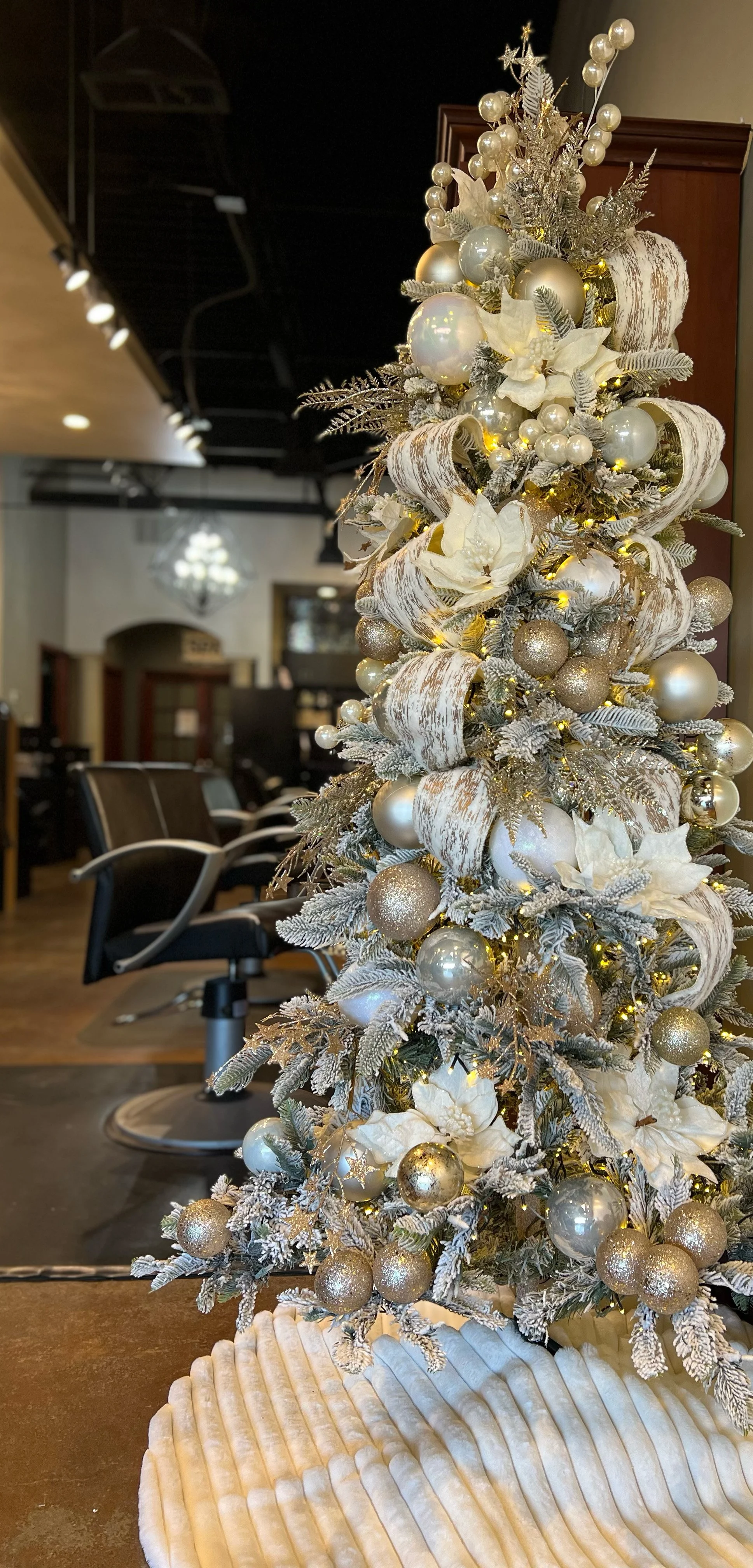 A small, decorative Christmas tree decorated with silver, white, and gold ornaments, ribbons, and lights, placed on a table in a salon or similar space.