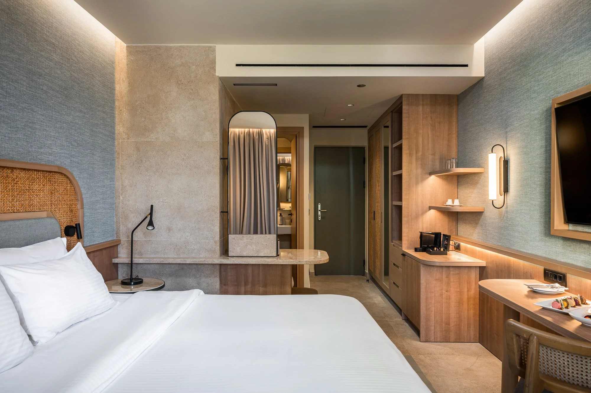 Modern hotel room with a bed, bedside lamp, small table, and a kitchenette area with shelves, a coffee maker, and a TV on the wall.