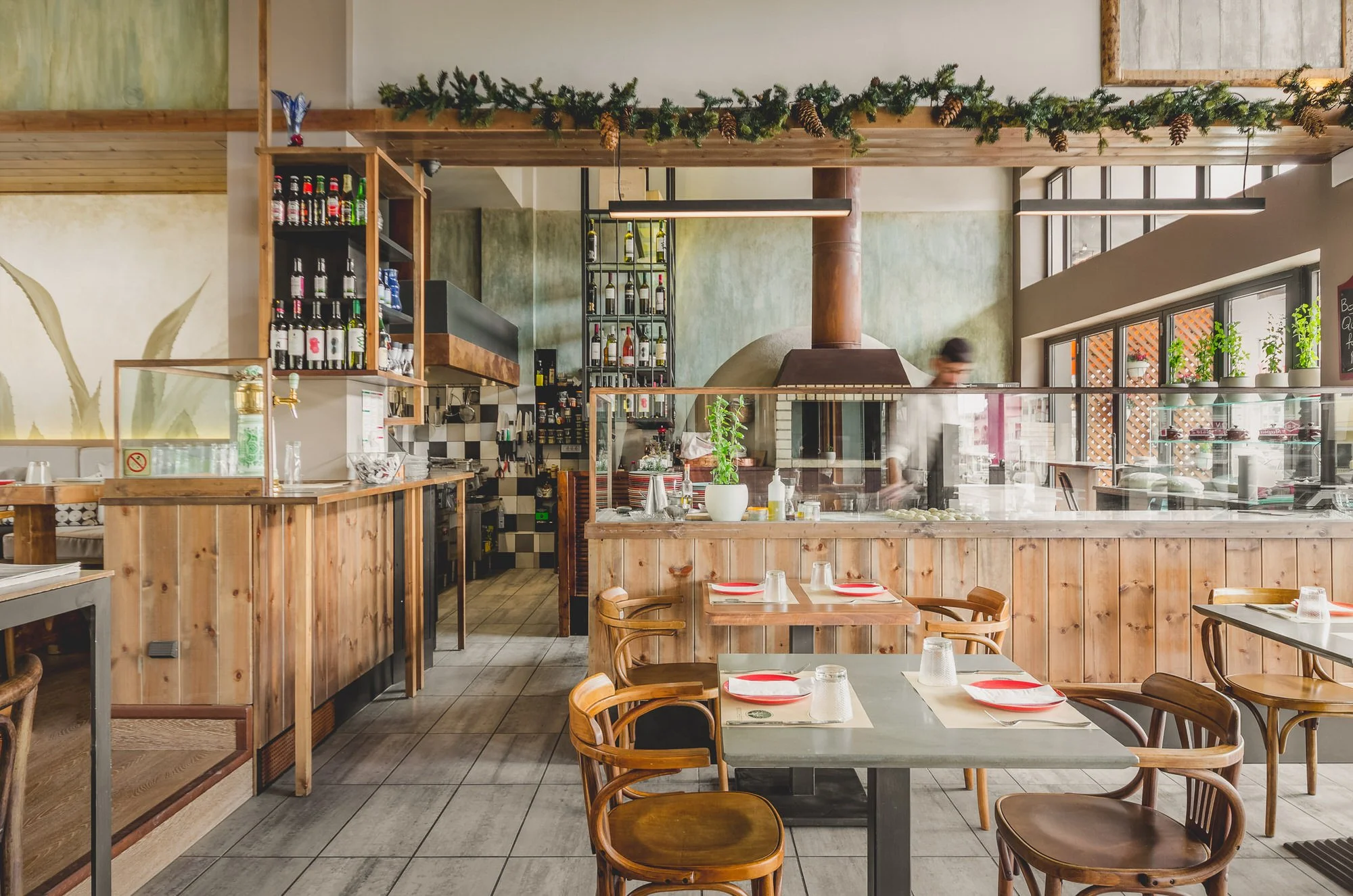 Interior of a cozy restaurant, a bar area with wine bottles on shelves, and a kitchen visible in the background with a chef working. Decor includes greenery and large windows with natural light. Hotels, Villas, Airbnb photography. Corfu, Greece.