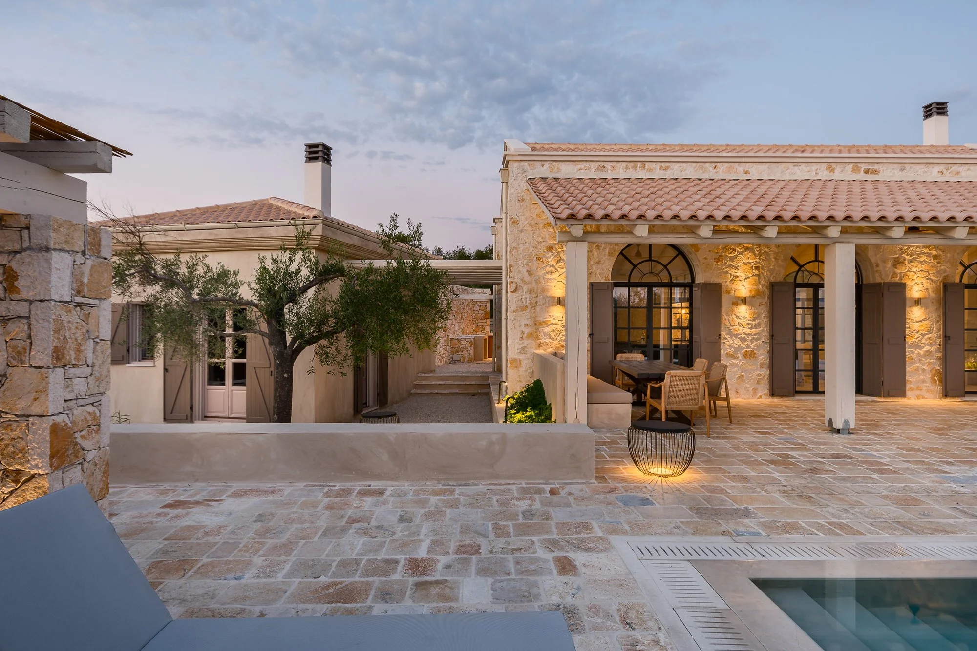A Mediterranean-style house with stone walls, arched windows, and a tiled roof, illuminated by warm outdoor lighting during twilight, featuring a patio area, a tree, and a glimpse of a swimming pool. Hotels, Villas, Airbnb photography. Corfu, Greece.