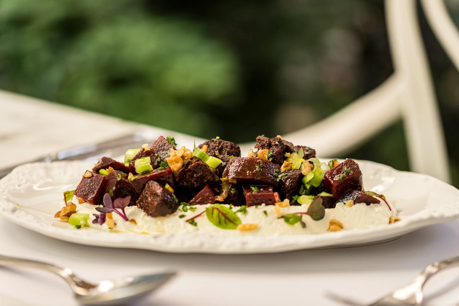 A white ornate plate with a dish of cooked beets, garnished with chopped green onions, microgreens, and nuts, set on a table with a silver spoon nearby. Hotels, Villas, Airbnb photography. Corfu, Greece.