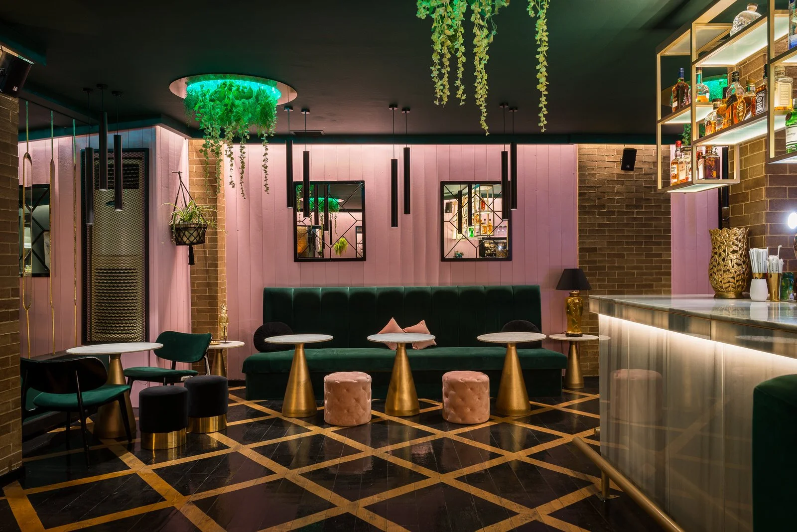Interior of a trendy bar or lounge with pink wooden walls, green velvet seating, gold accent tables, hanging plants, and illuminated bottles on shelves. Hotels, Villas, Airbnb photography. Corfu, Greece.