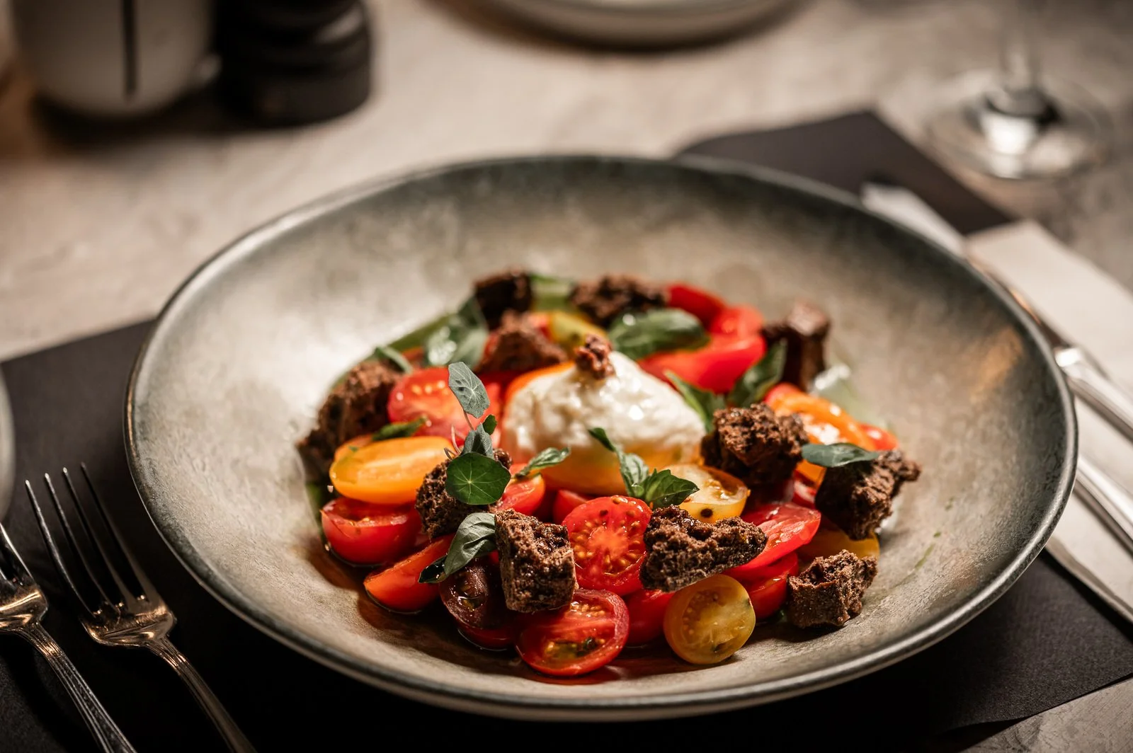 A salad with cherry tomatoes, beef crumble, a dollop of white cheese or cream, and fresh green herbs in a gray bowl on a black placemat with silverware. Hotels, Villas, Airbnb photography. Corfu, Greece.