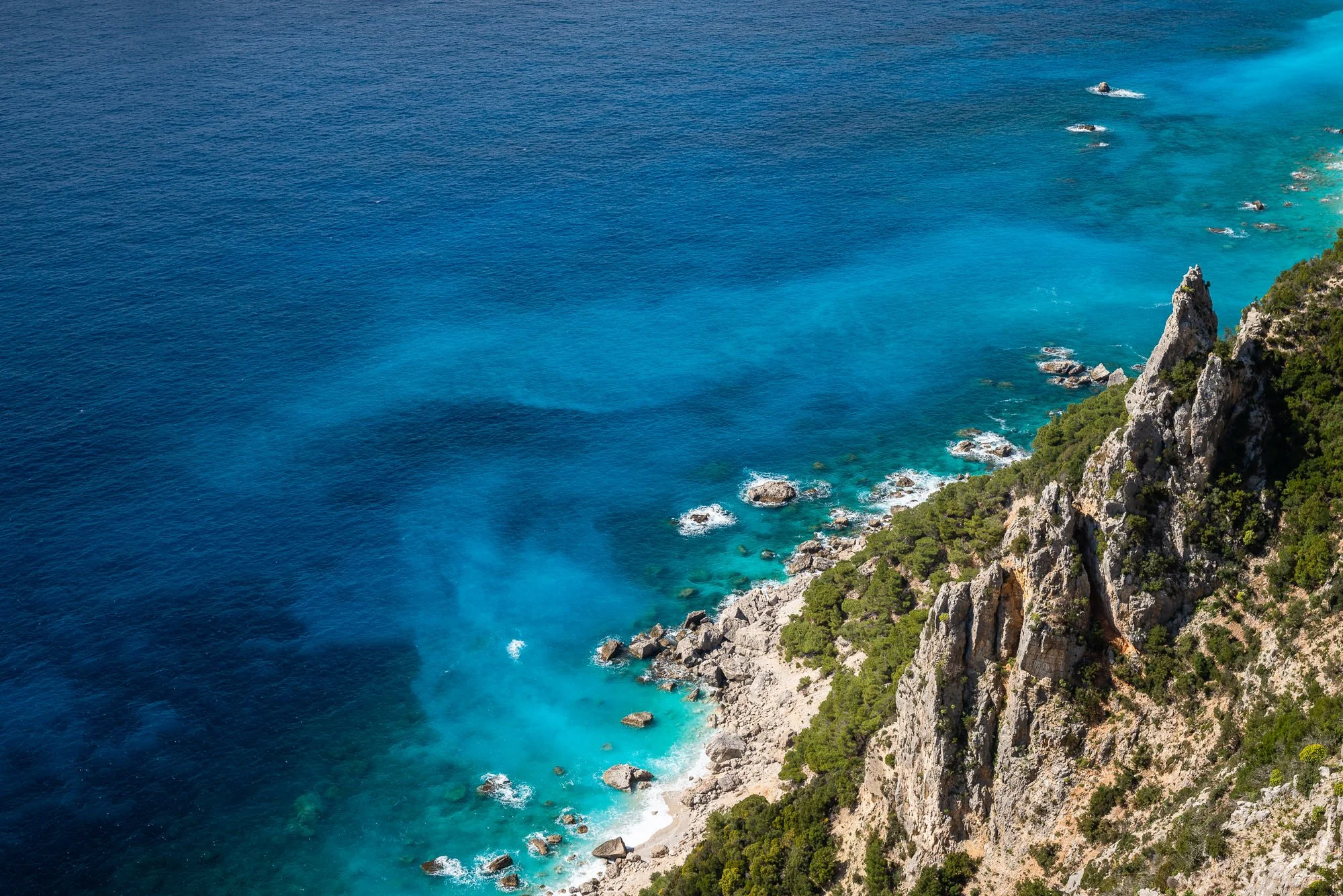 Aerial view of a rocky coastline with green shrubbery, turquoise water near the shore, and deeper blue ocean further out. Hotels, Villas, Airbnb photography. Corfu, Greece.