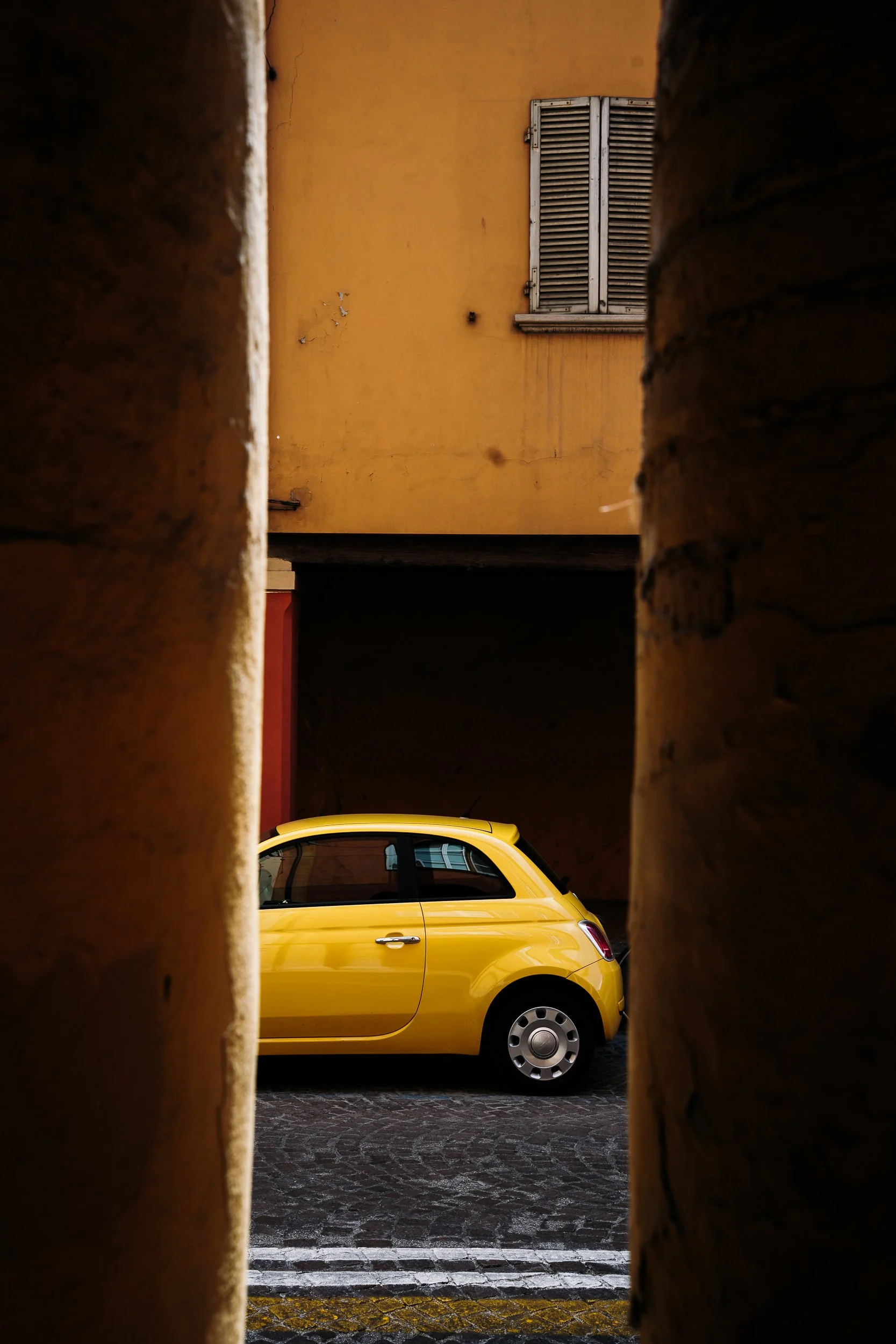 A yellow compact car parked on a cobblestone street, viewed through a narrow opening between two brick walls, with a yellow building and a window with closed shutters in the background.