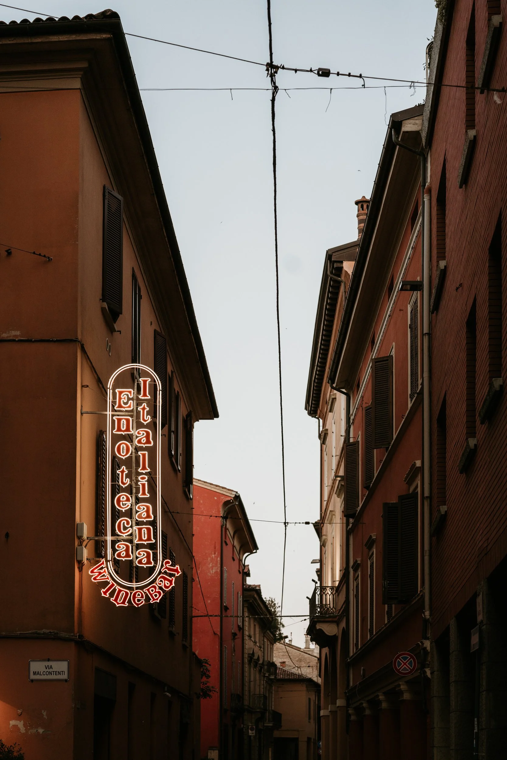 Narrow street between old buildings in Italy with a neon sign reading 'Italiana Edotecca' on the left side.