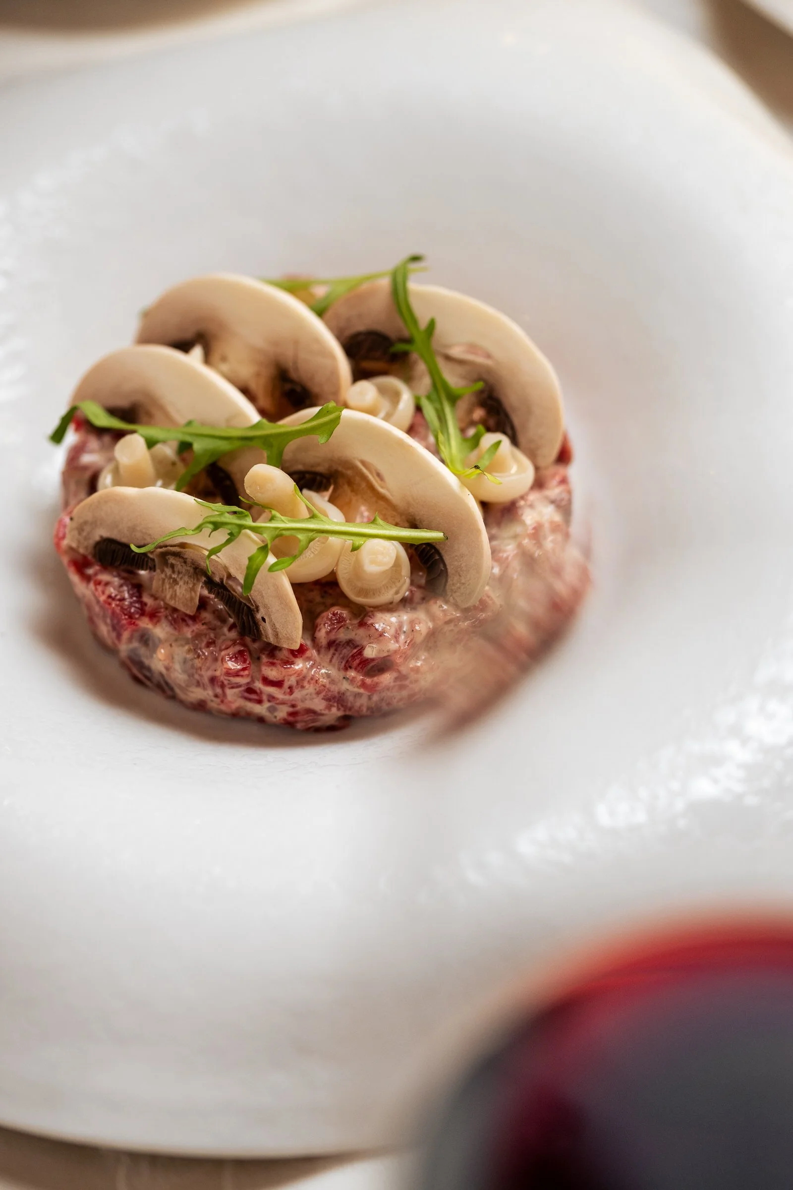 Close-up of a raw beef steak topped with sliced white mushrooms, small white onions, and garnished with a sprig of arugula, served on a white plate.Hotels, Villas, Airbnb photography. Corfu, Greece.