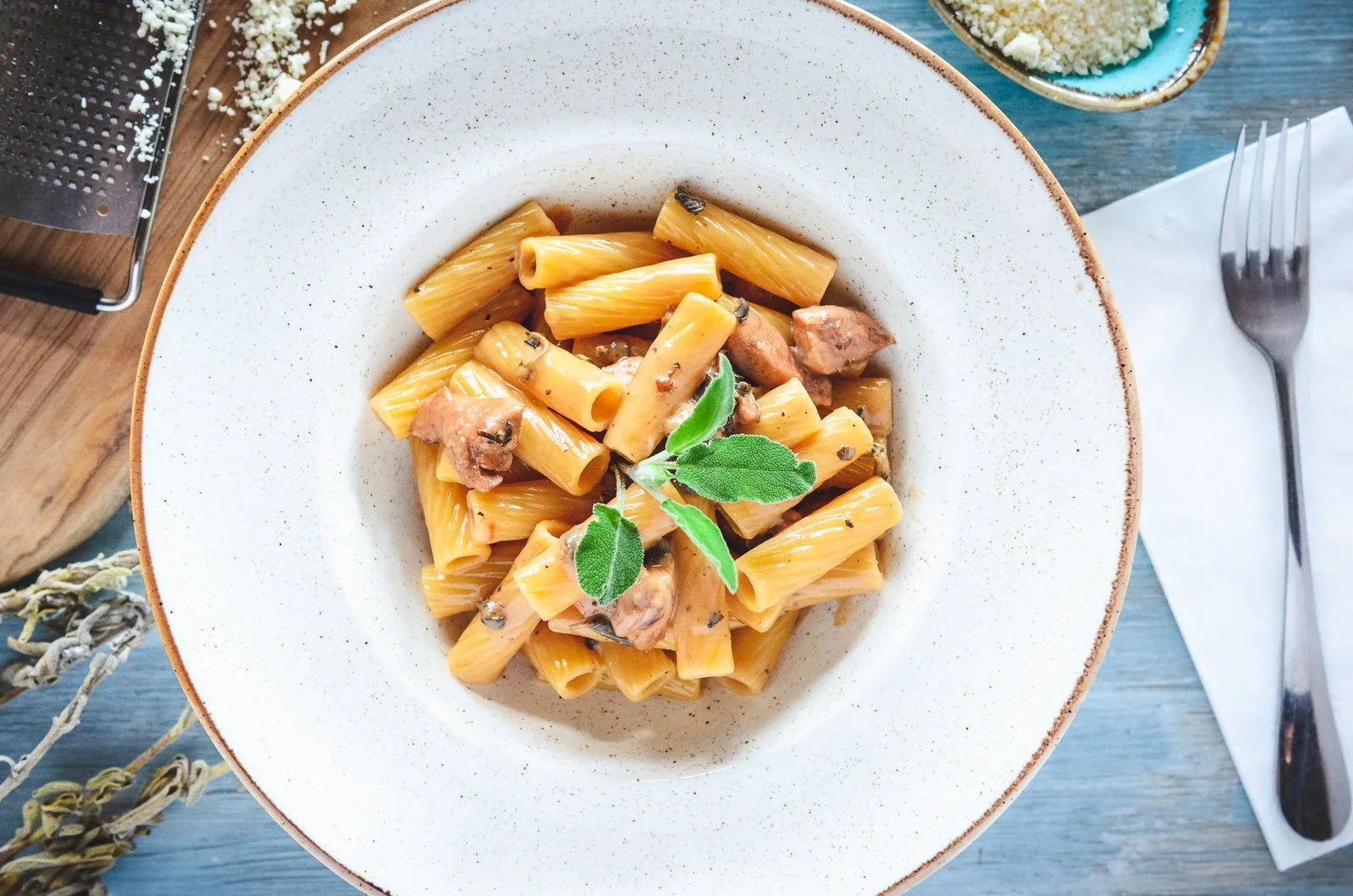 A plate of pasta with a creamy sauce, garnished with fresh sage leaves, on a blue wooden table with a fork, a napkin, and a small bowl of grated cheese nearby.Hotels, Villas, Airbnb photography. Corfu, Greece.