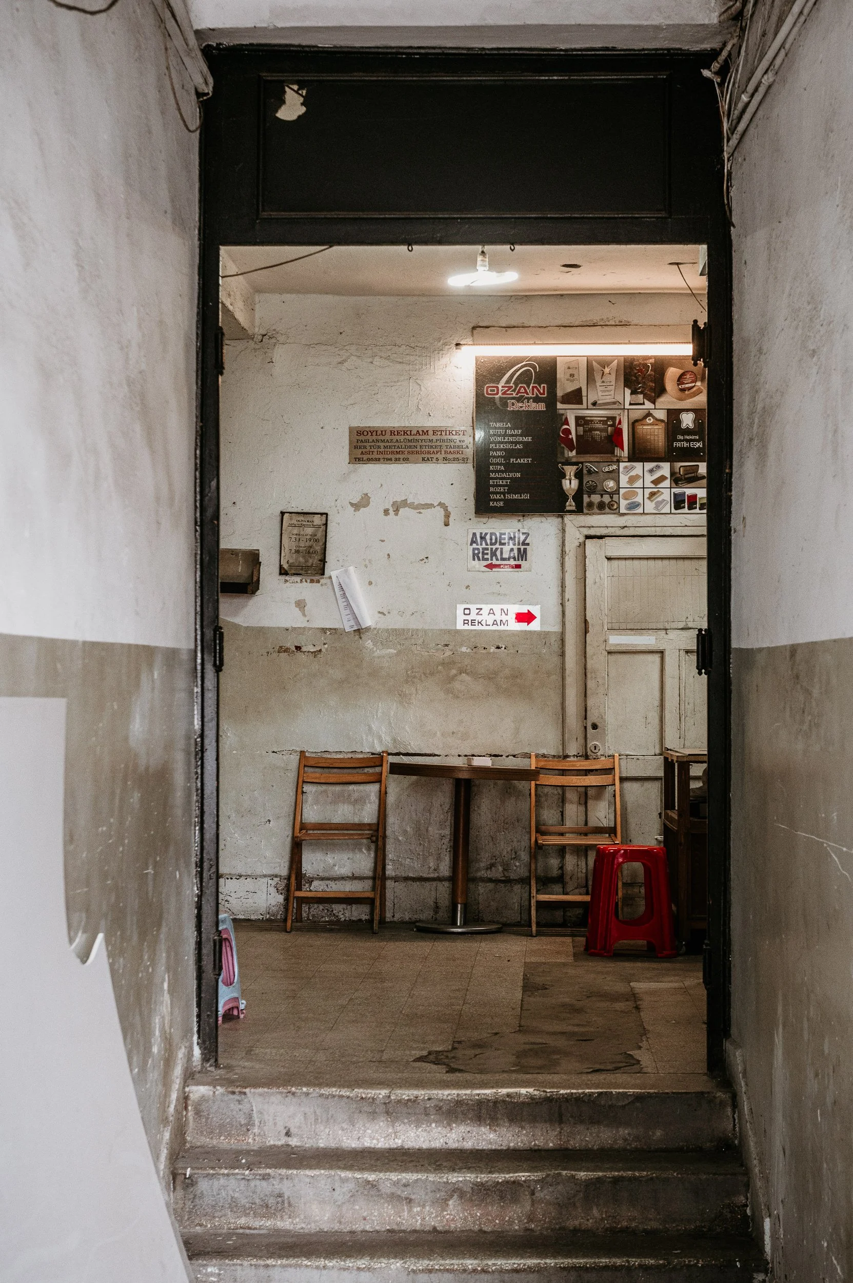Interior of an old, slightly worn room with two wooden chairs, a small red plastic stool, and a tabletop. The walls are weathered and painted in a cream color, and there are various signs and posters on the back wall, some with text in Turkish. There