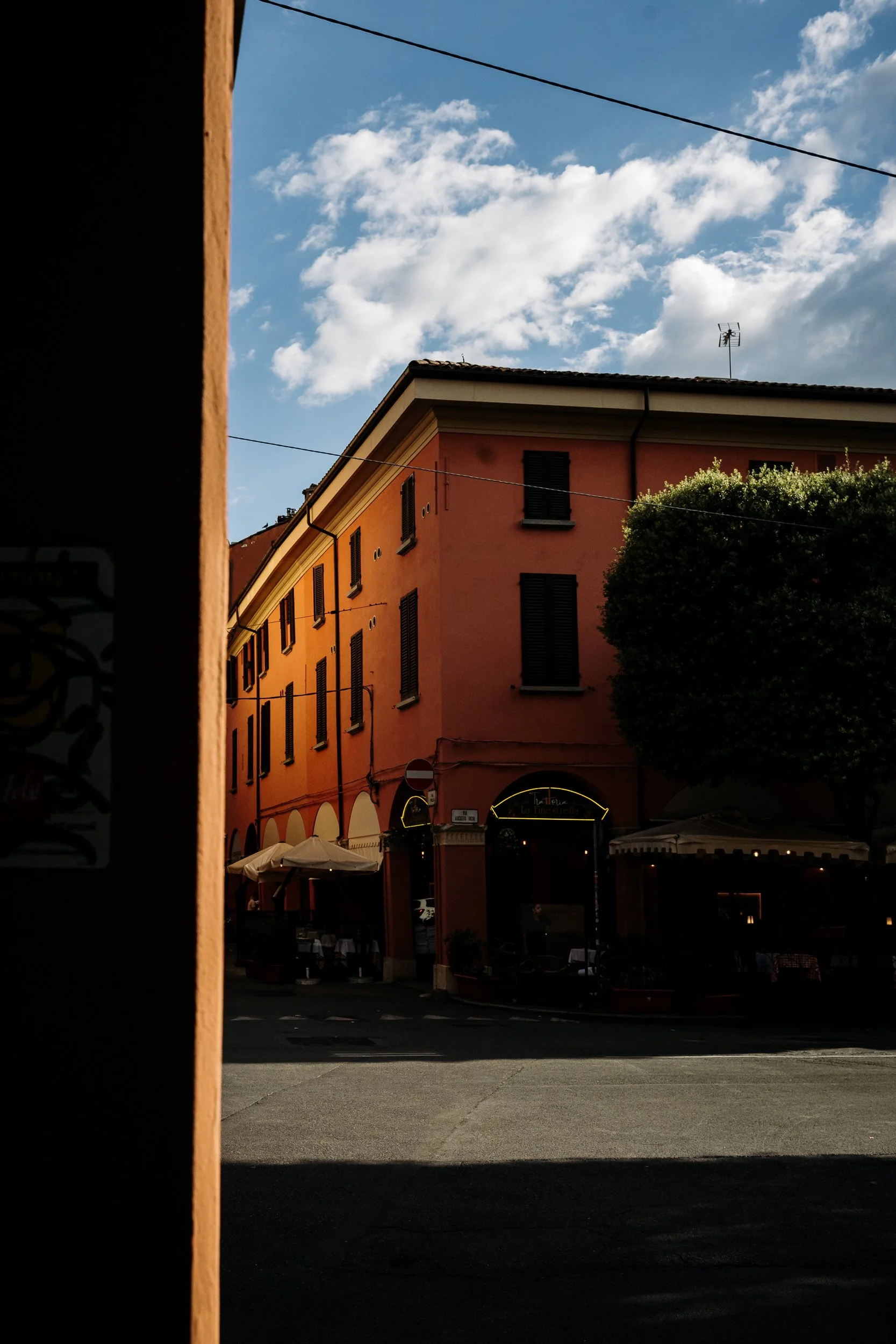 A street scene with a pink building, outdoor seating with umbrellas, a large tree, and a partly cloudy sky.