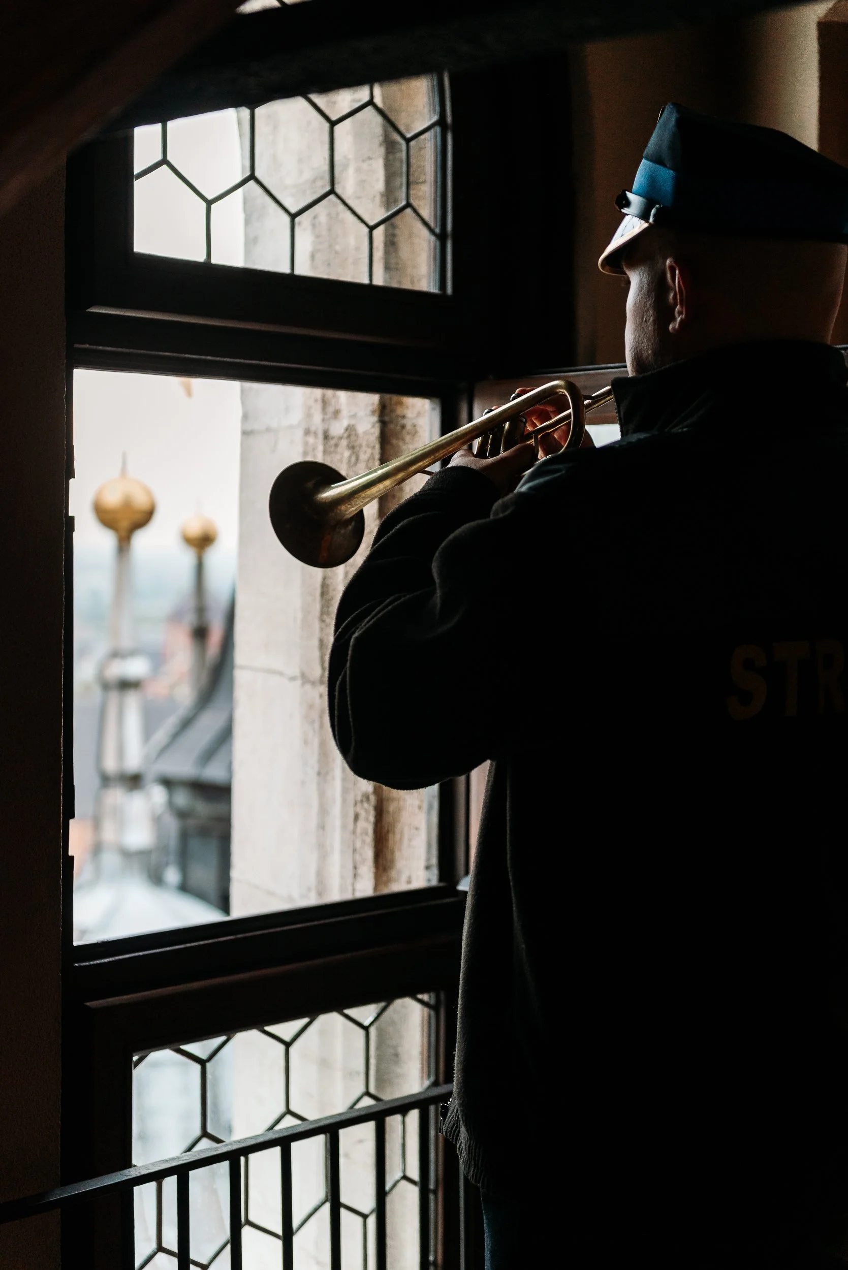 A person dressed in black, wearing a police or military cap, playing a brass trumpet near a window with stained glass and stone framework.
