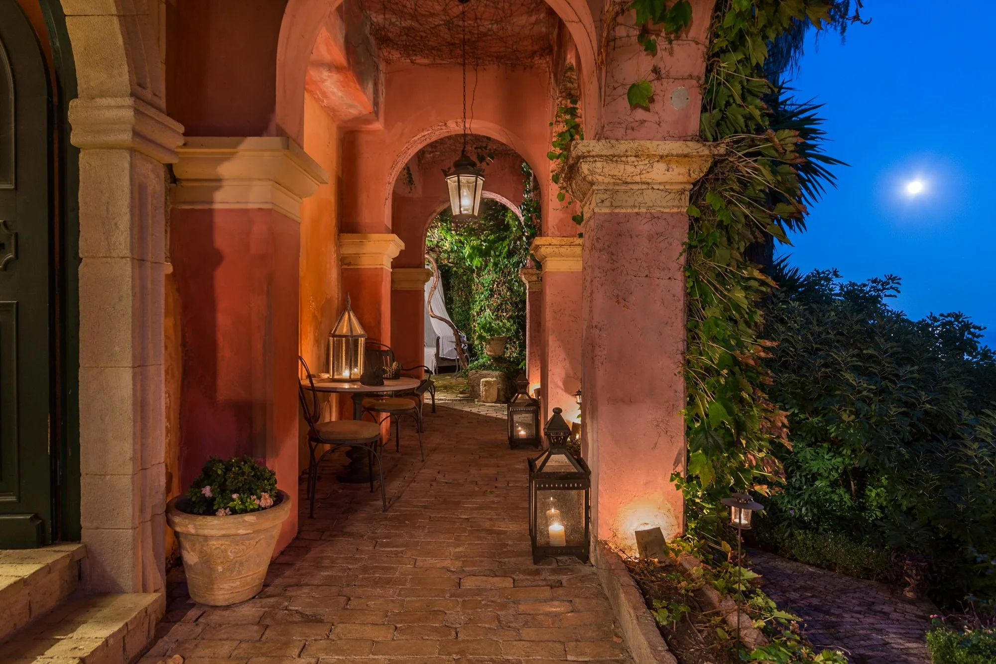 A nighttime scene of an outdoor patio with pink stucco walls, decorated with lanterns and candles, winding brick pathway, and lush greenery under a clear evening sky with a visible moon. Hotels, Villas, Airbnb photography. Corfu, Greece.