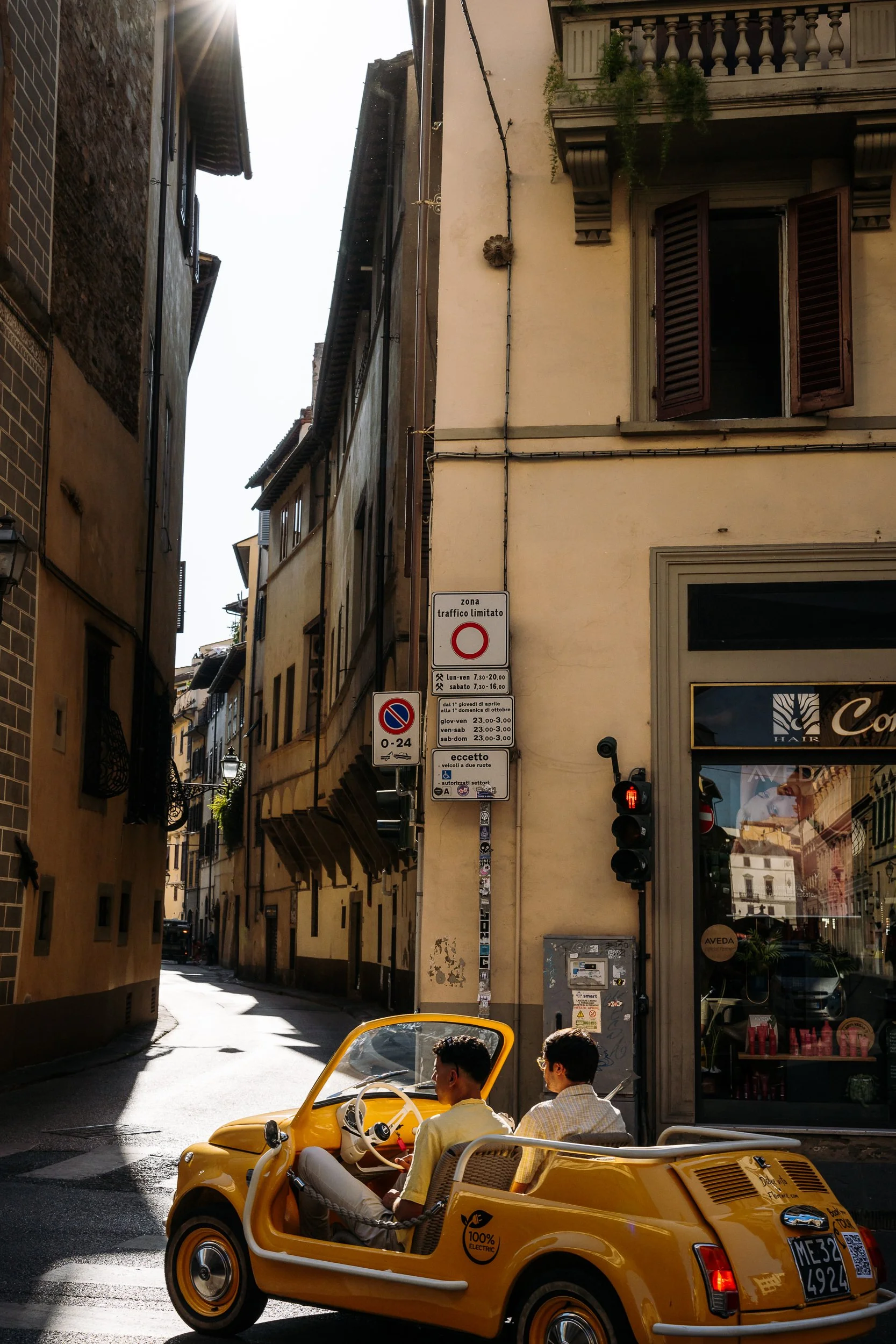 Two young men in a yellow electric vintage-style car driving through a narrow European street with old buildings and signs.