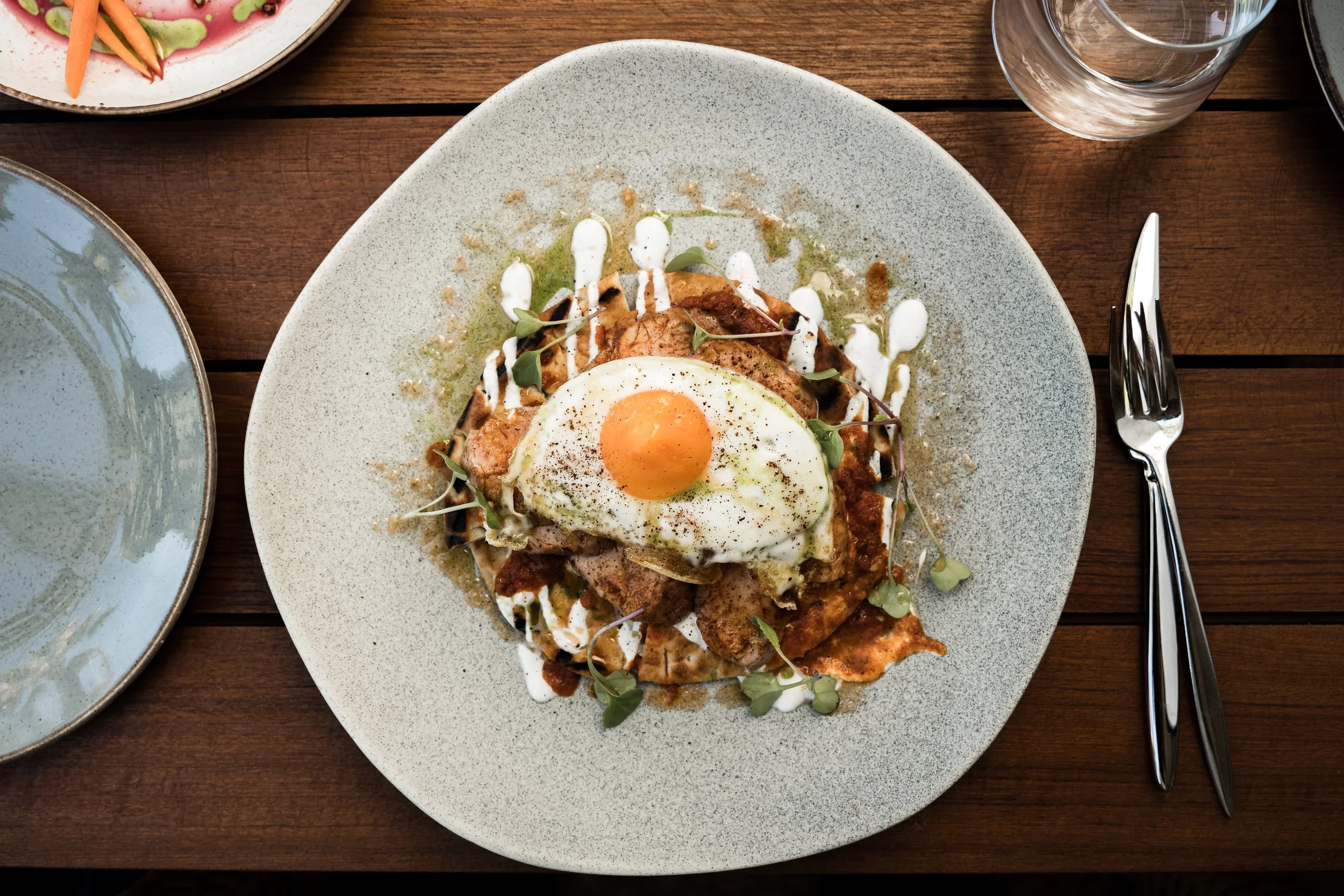 A plate of food with a fried egg on top, garnished with microgreens, served on a bed of meat, sauce, and garnishes, on a wooden table with a glass of water and cutlery nearby.Hotels, Villas, Airbnb photography. Corfu, Greece.