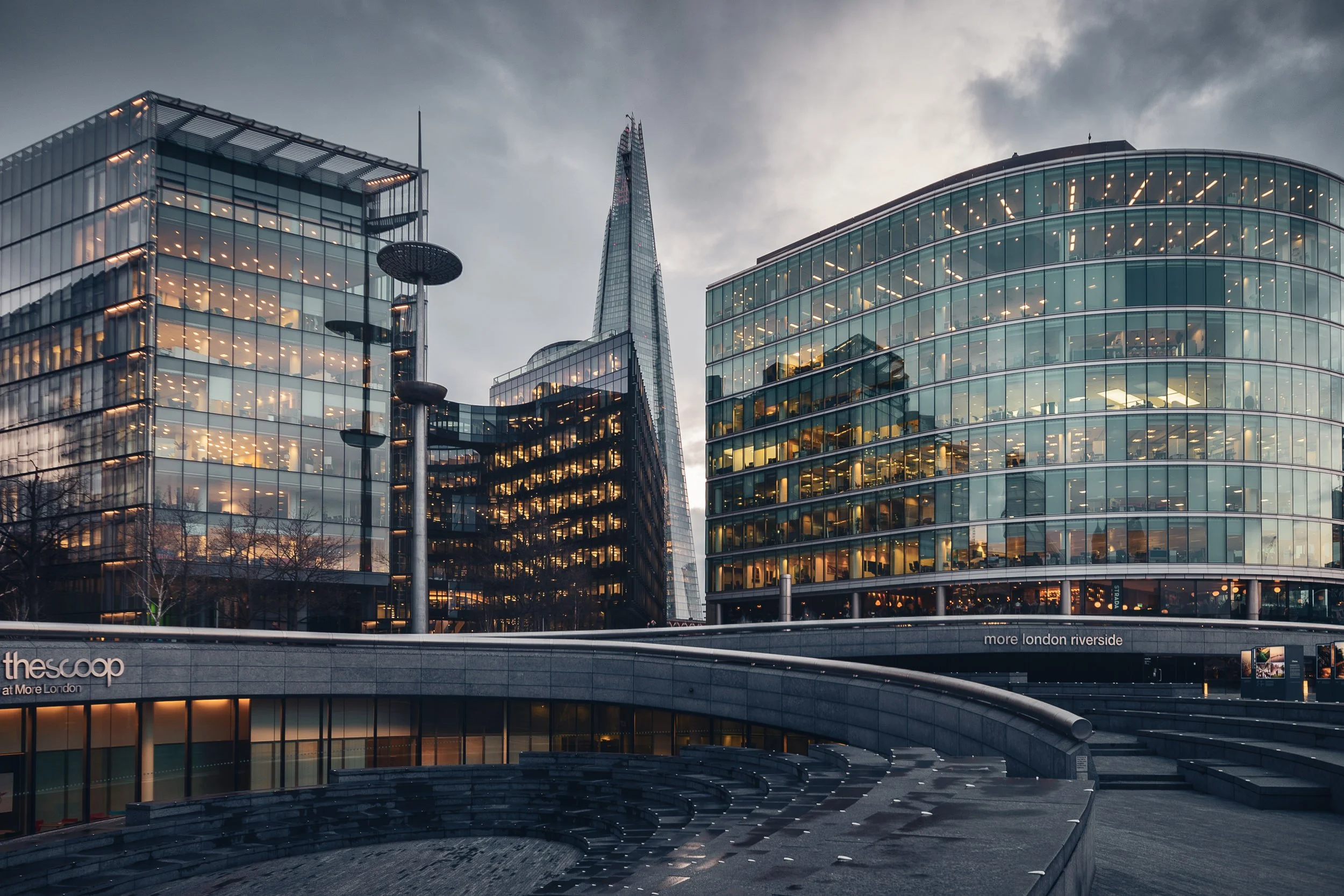Modern glass buildings in London's riverfront area during dusk, with a cloudy sky overhead.