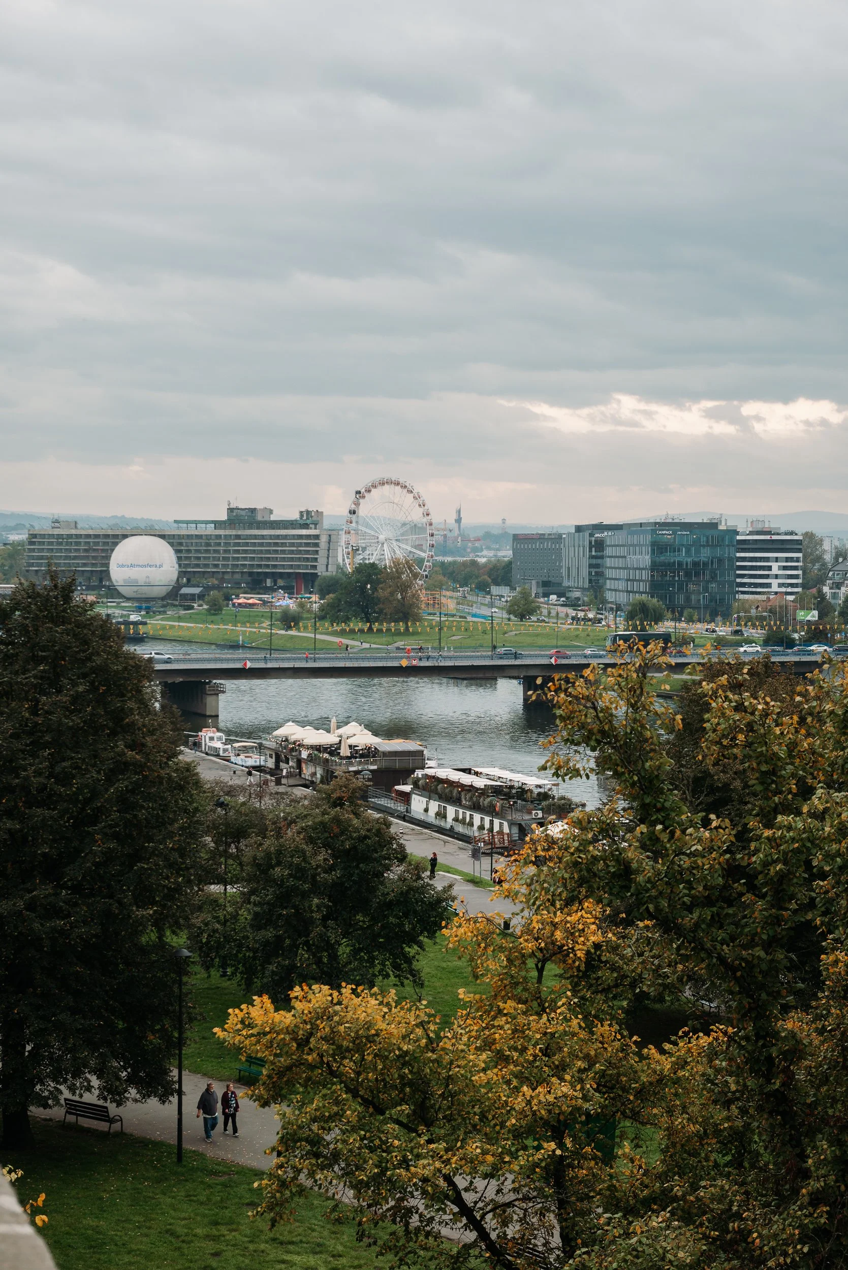 A cityscape with a river, trees with autumn foliage, a bridge, buildings, a ferris wheel, and a large white spherical structure under a cloudy sky.