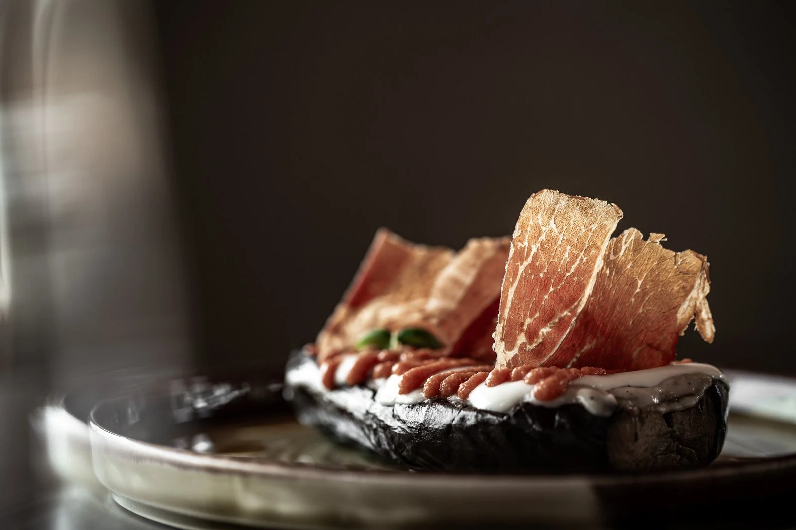 Close-up of an appetizer with crispy prosciutto, creamy cheese, and fresh herbs on a dark plate, with a blurred dark background. Hotels, Villas, Airbnb photography. Corfu, Greece.