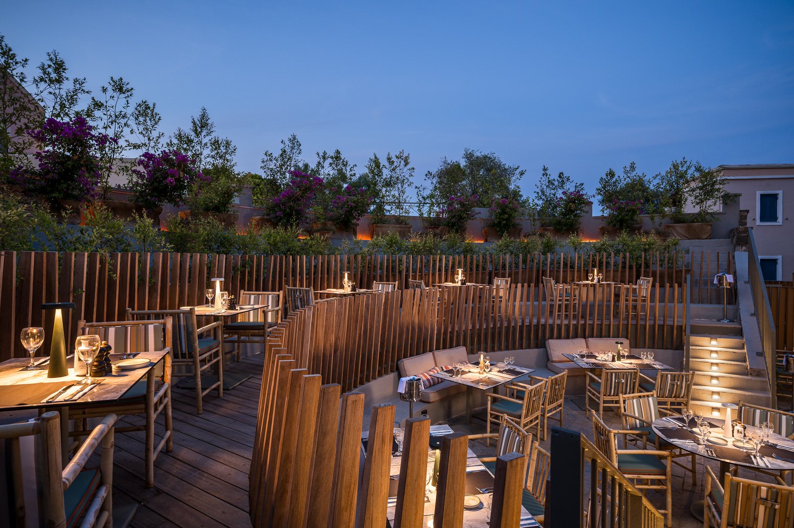 Outdoor restaurant patio with wooden tables and chairs, set for dinner with glasses, silverware, and candles, surrounded by a wooden fence and potted plants against a blue evening sky. Hotels, Villas, Airbnb photography. Corfu, Greece.