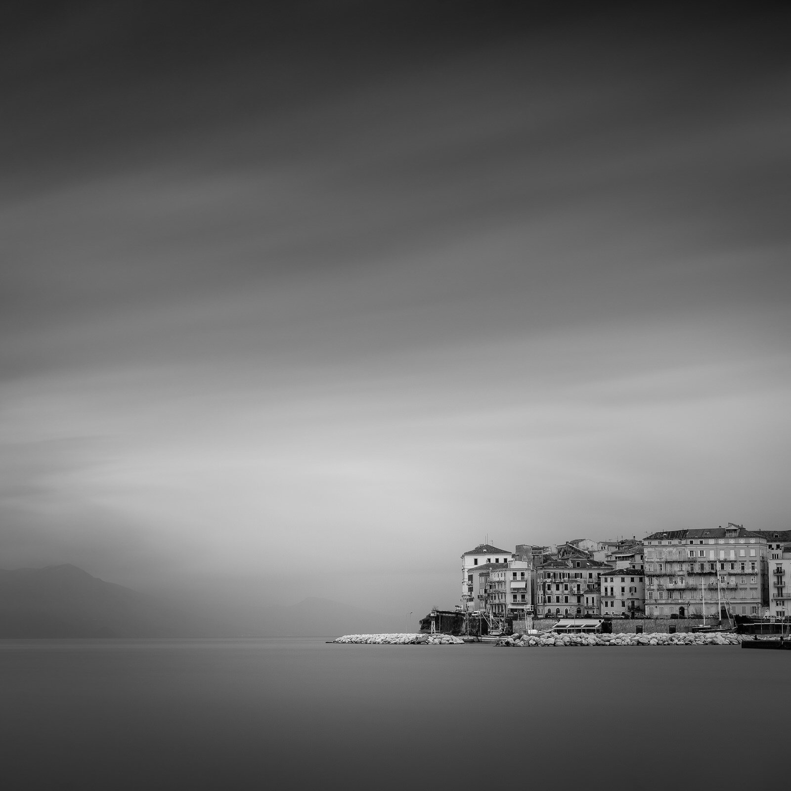 Black and white photo of a coastal town with buildings along the shoreline, calm water in the foreground, and cloudy sky above.