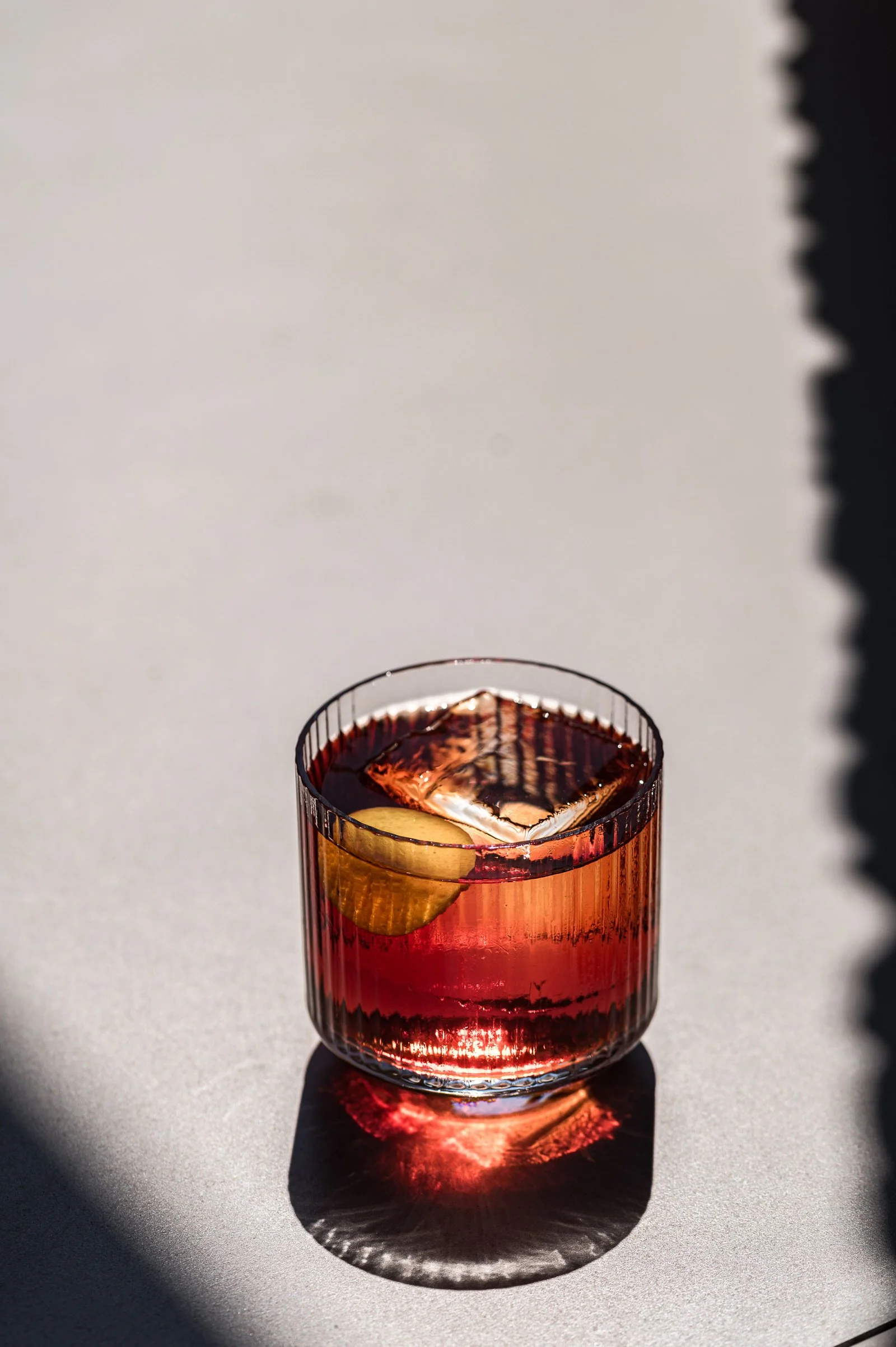 A glass of red or dark pink cocktail with a lemon wedge and ice, placed on a light-colored surface with shadows and reflections.Hotels, Villas, Airbnb photography. Corfu, Greece.