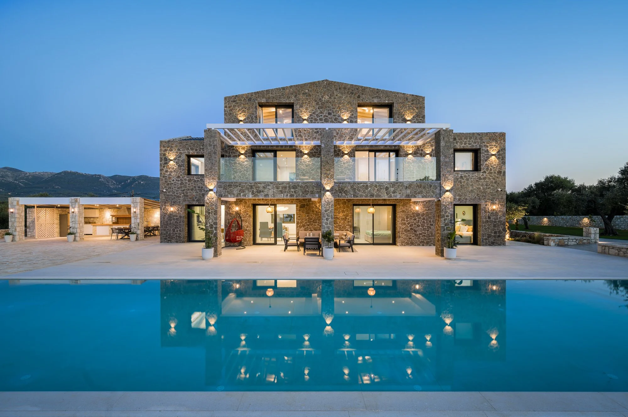 Luxury modern stone house with large outdoor patio, glass doors, and a swimming pool, set against mountain backdrop at dusk. Hotels, Villas, Airbnb photography. Corfu, Greece.