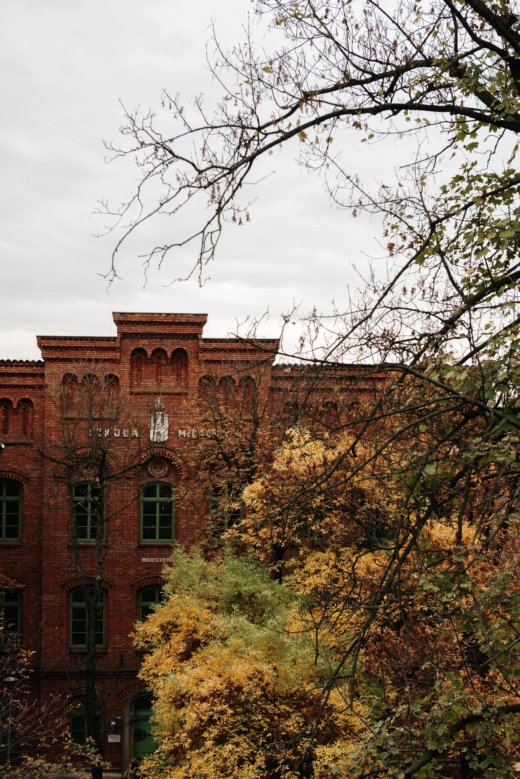 A red brick building partially obscured by autumn-colored trees and branches, with a cloudy sky overhead.