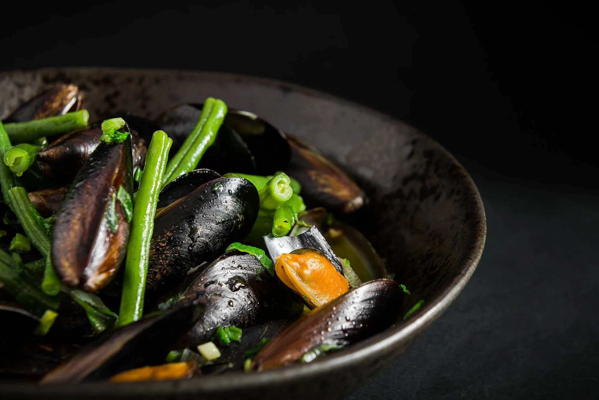 A black bowl filled with cooked mussels, green beans, and chopped green onions on a dark background. Hotels, Villas, Airbnb photography. Corfu, Greece.